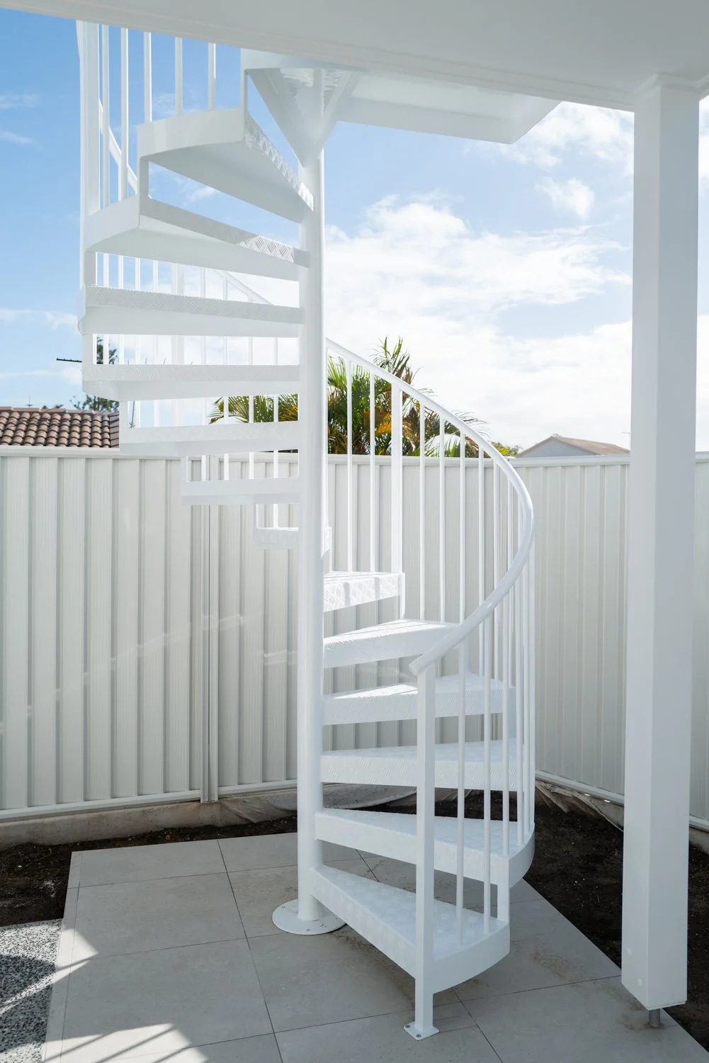 A white outdoor spiral staircase with metal railings, leading to an upper level, outside a house with a white fence and a partly cloudy sky.
