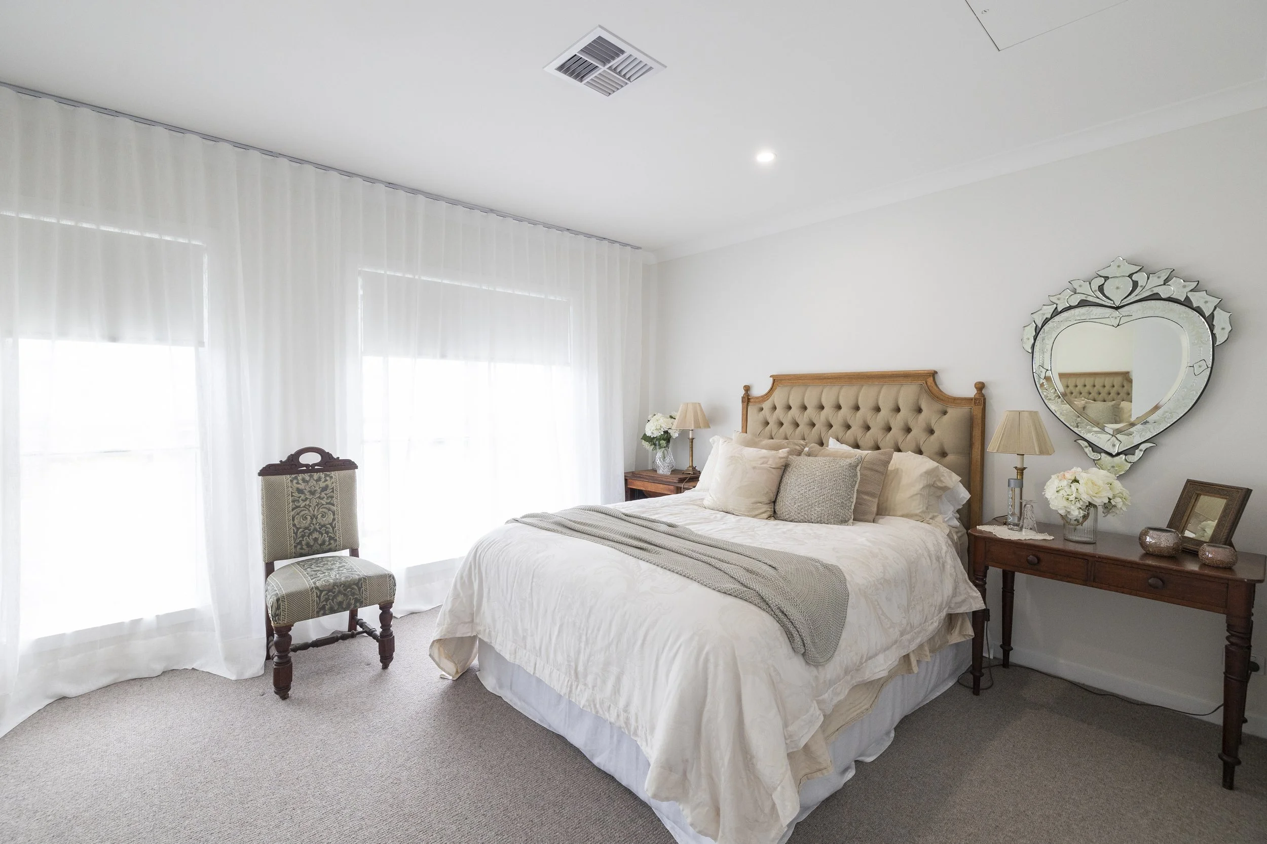 A cozy bedroom with a tufted beige headboard, white bedding, and beige accent pillows. A wooden side table with a lamp, flowers, and framed photos is beside the bed. A decorative mirror hangs on the wall, and sheer white curtains cover the windows. A