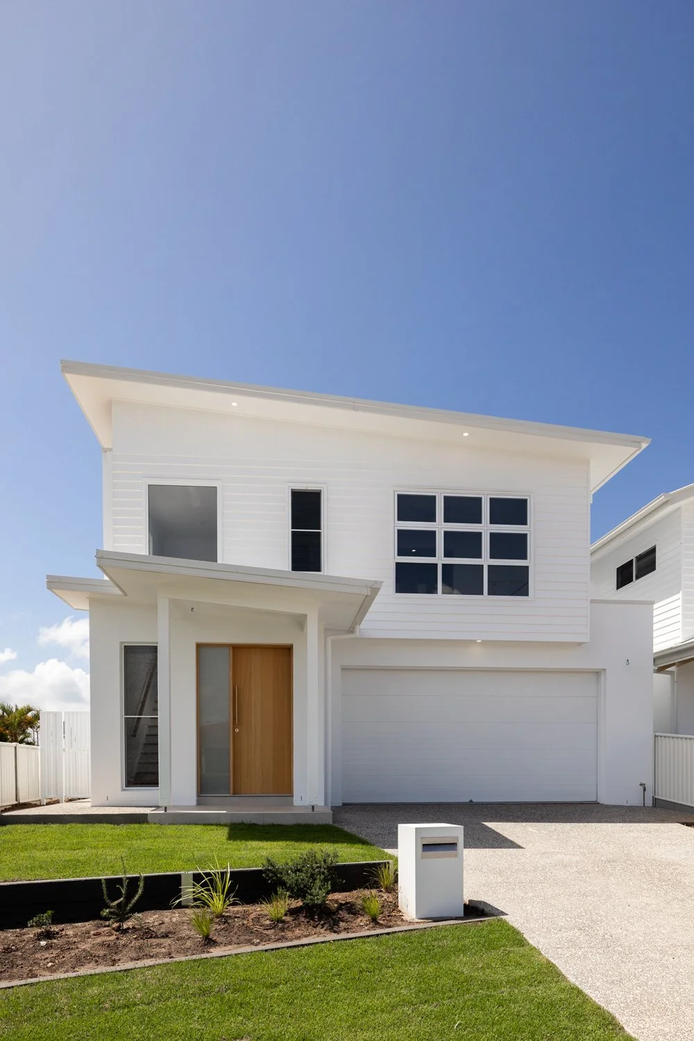 Modern white two-story house with a wooden front door, large windows, a white garage door, and a front lawn with plants, under a clear blue sky.