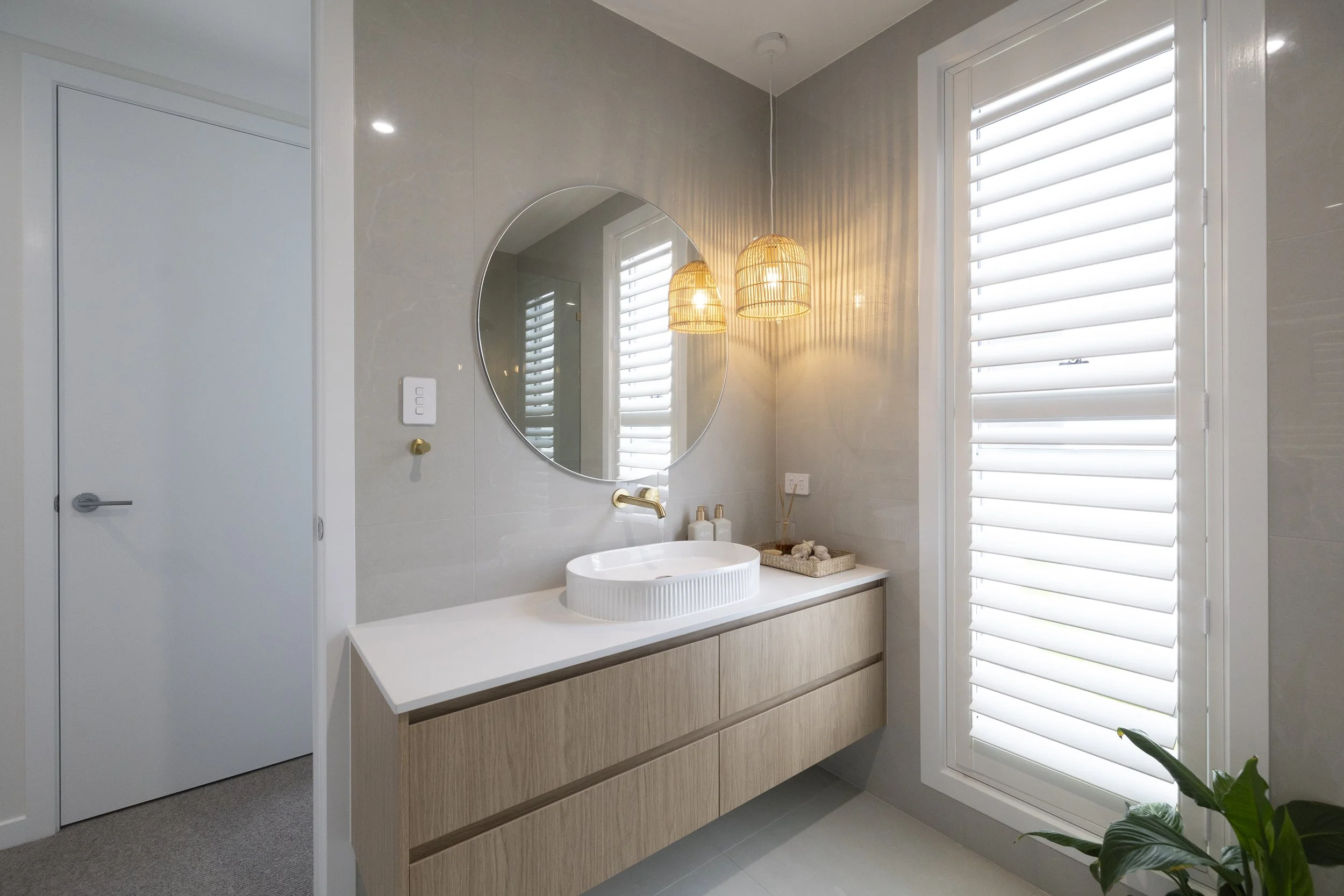Modern bathroom with a round mirror, woven pendant lights, a white countertop with a vessel sink, and large windows with white plantation shutters.