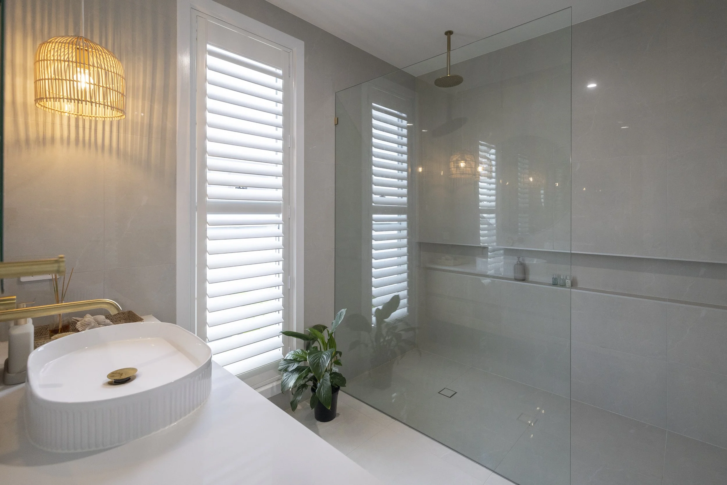 Modern bathroom with white walls, large window with white shutters, a glass shower enclosure, a white sink with a gold faucet, a small potted plant, and a wall-mounted wooden light fixture.