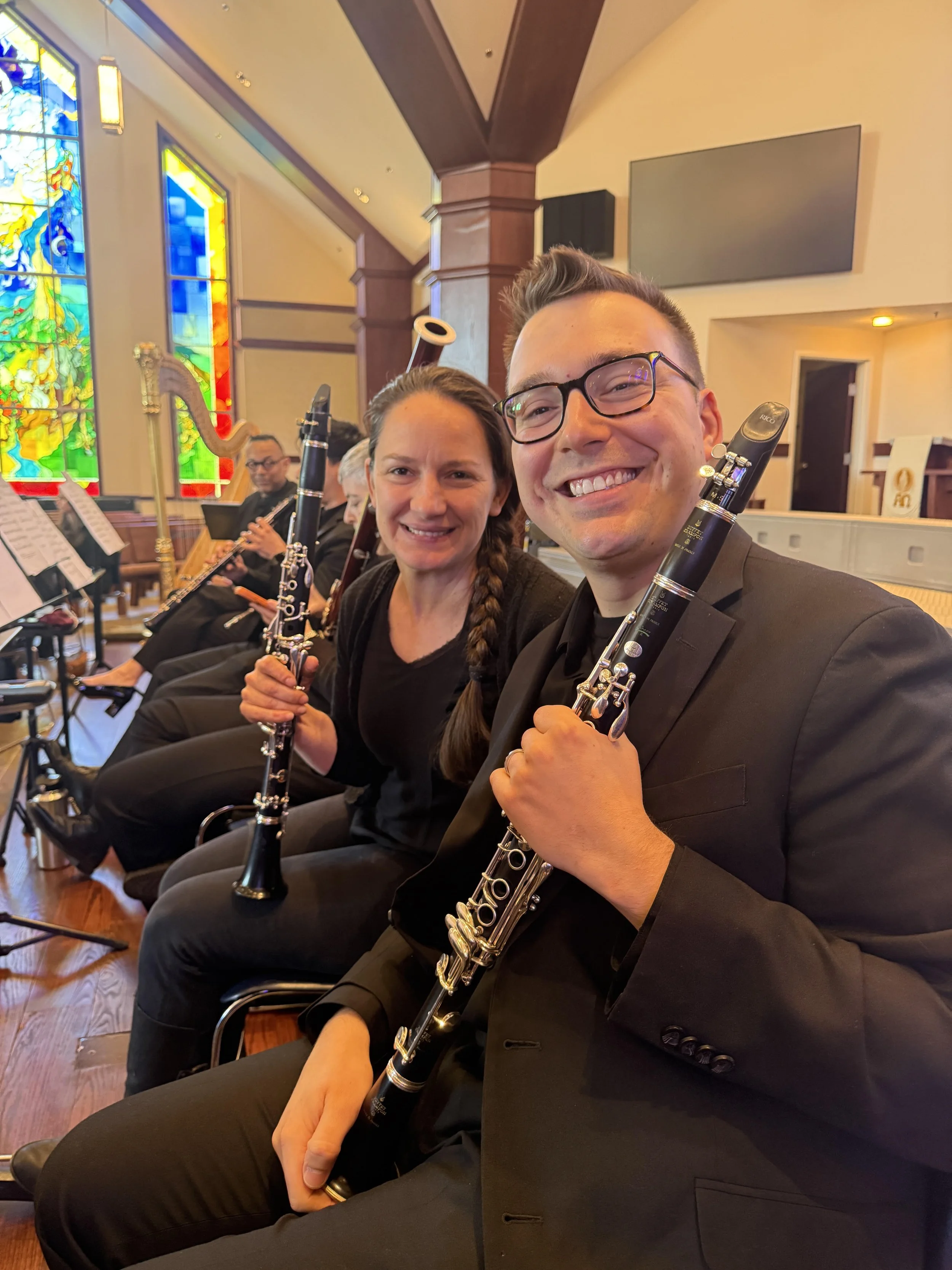 Two clarinet players, holding clarinets, smiling for a selfie with stained glass windows and other musicians in the background.