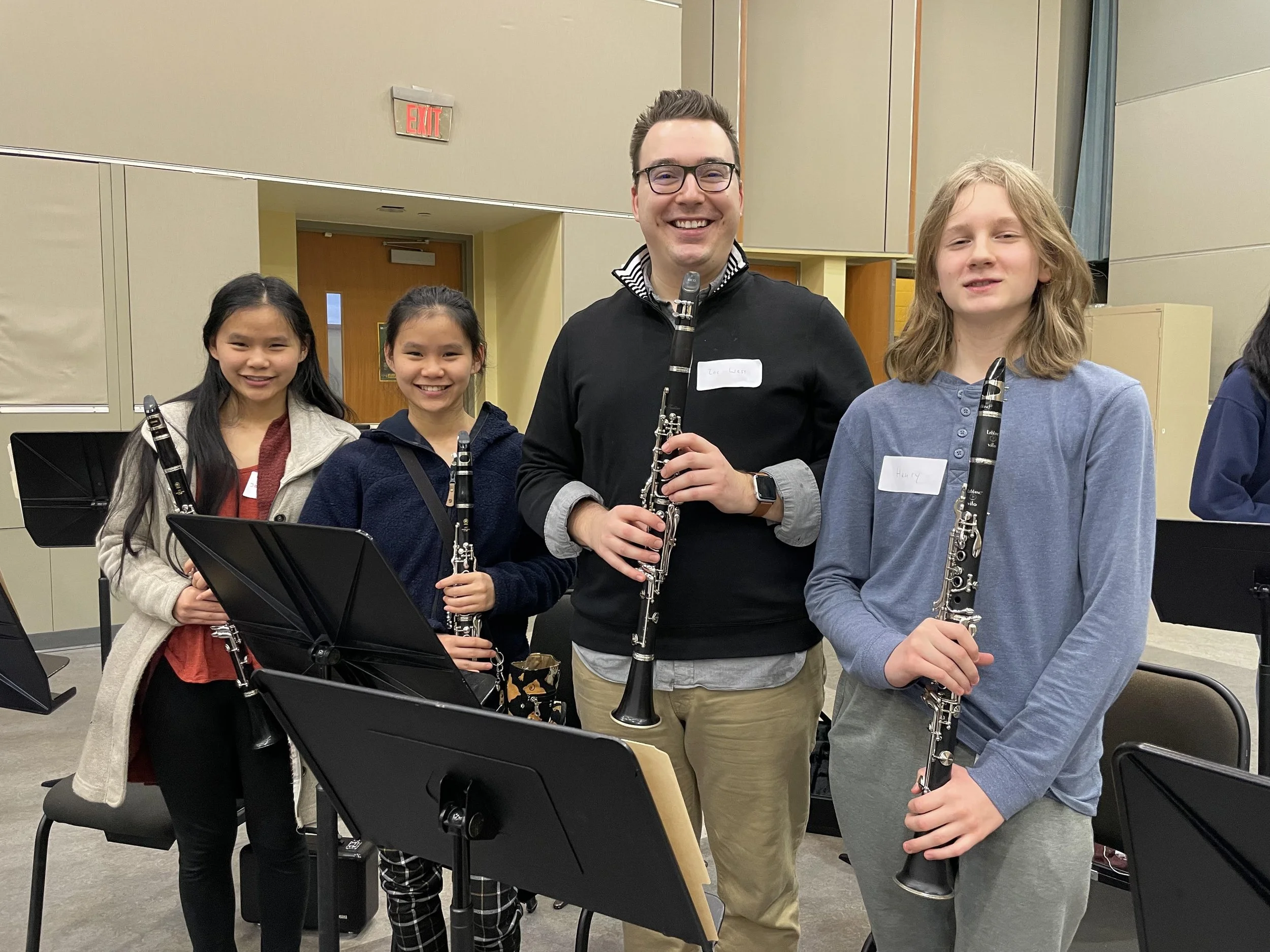 Four children and an adult man holding clarinets, standing in a music rehearsal room with music stands and a beige wall in the background.