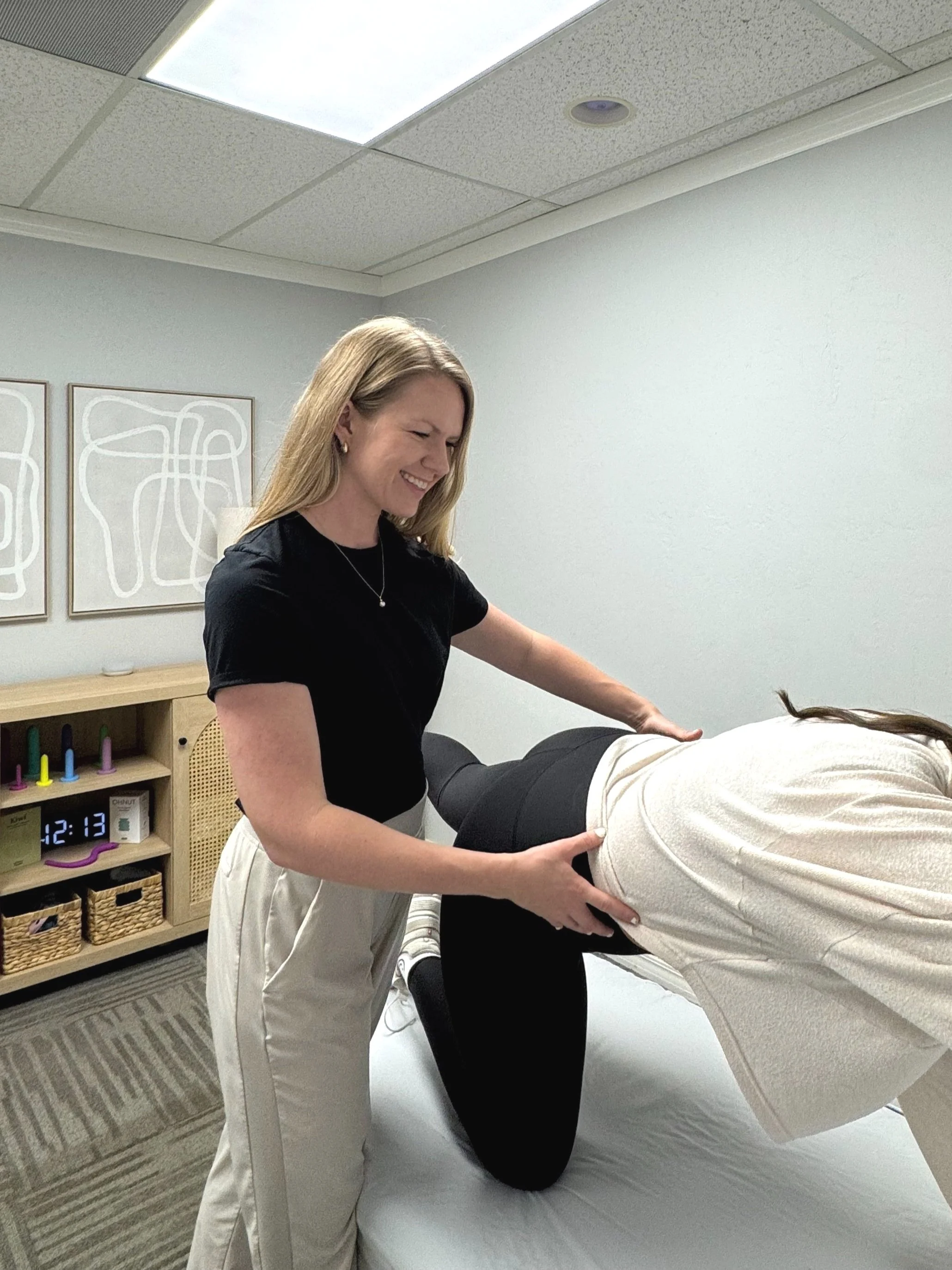 A woman assisting another person with a physical therapy exercise in a clinical room with white walls and abstract art.