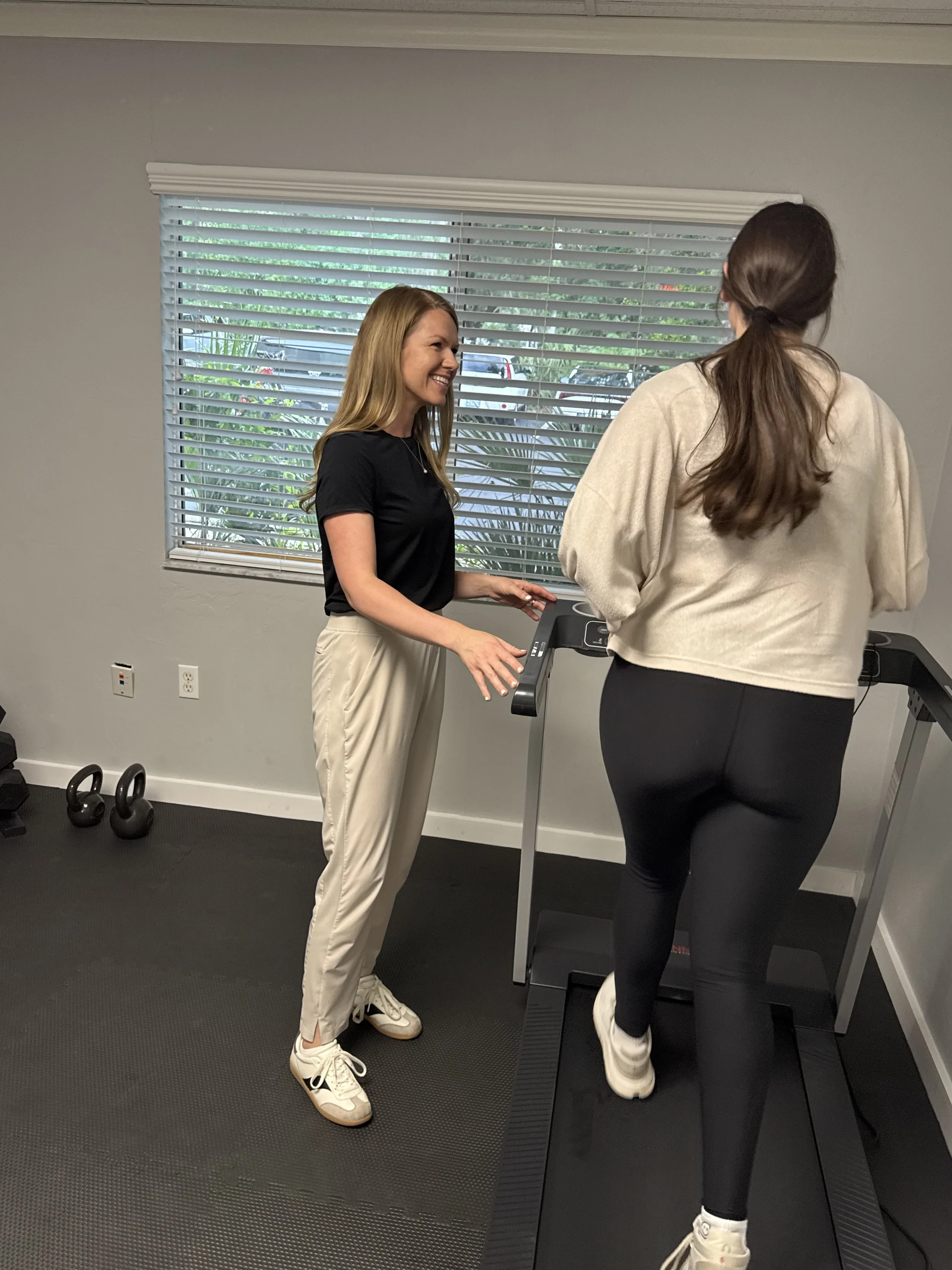 A woman on a treadmill and a woman standing next to her in a gym, smiling and talking.