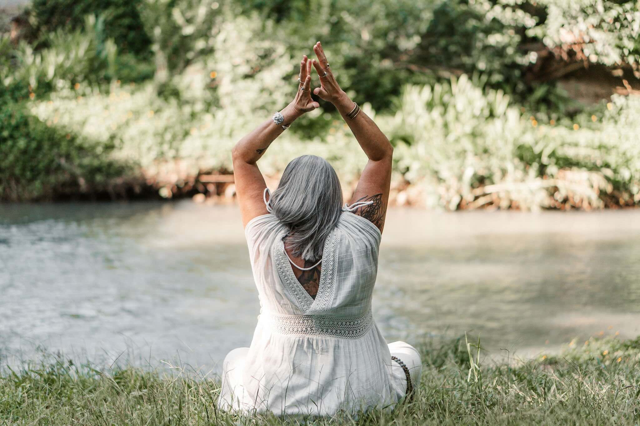An elderly woman with gray hair and tattoos, sitting cross-legged on grass beside a river, facing away, with arms raised and hands pressed together in a yoga pose.