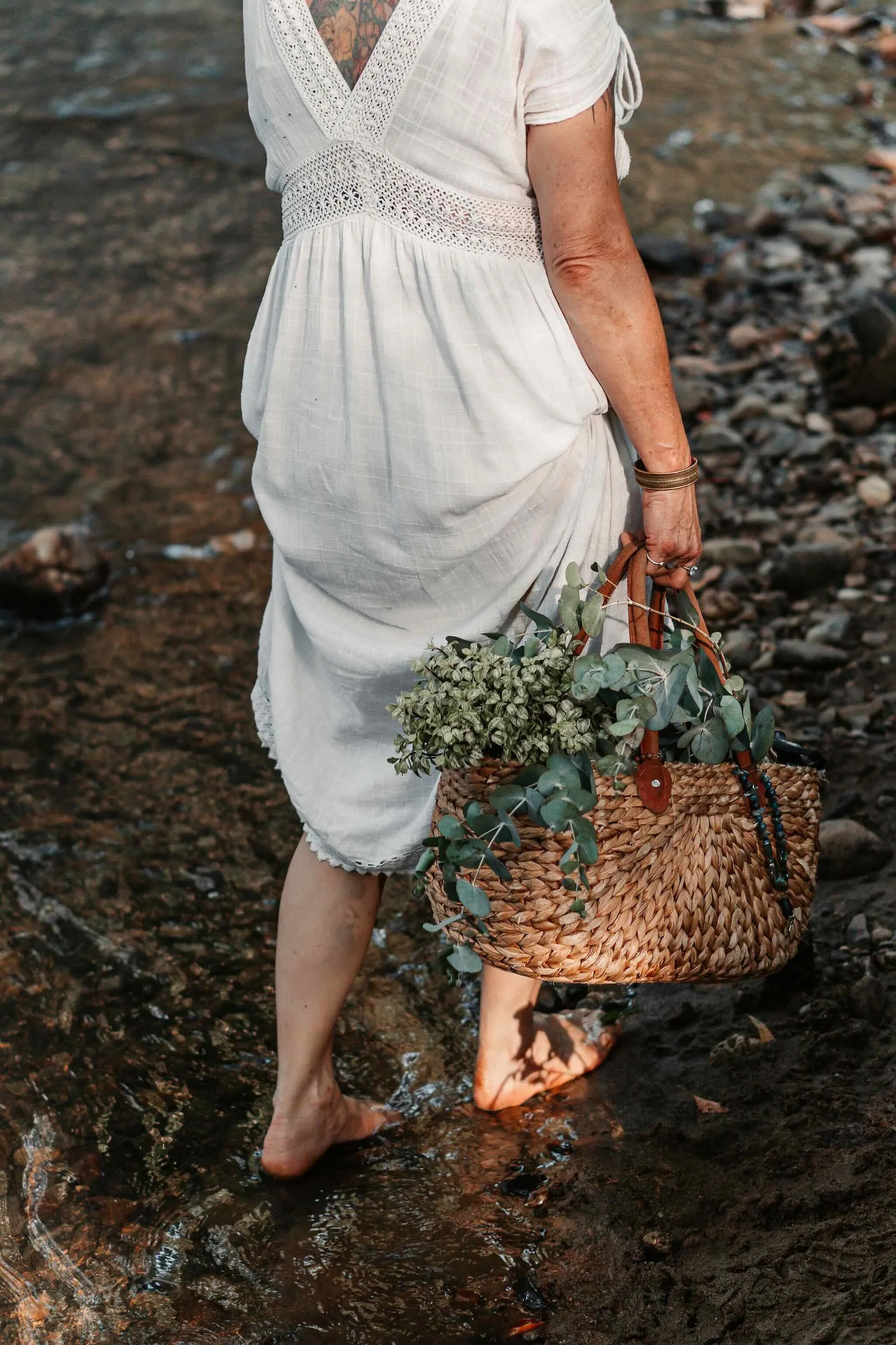 A woman in a white dress walking on a rocky shore, carrying a woven basket filled with greenery.