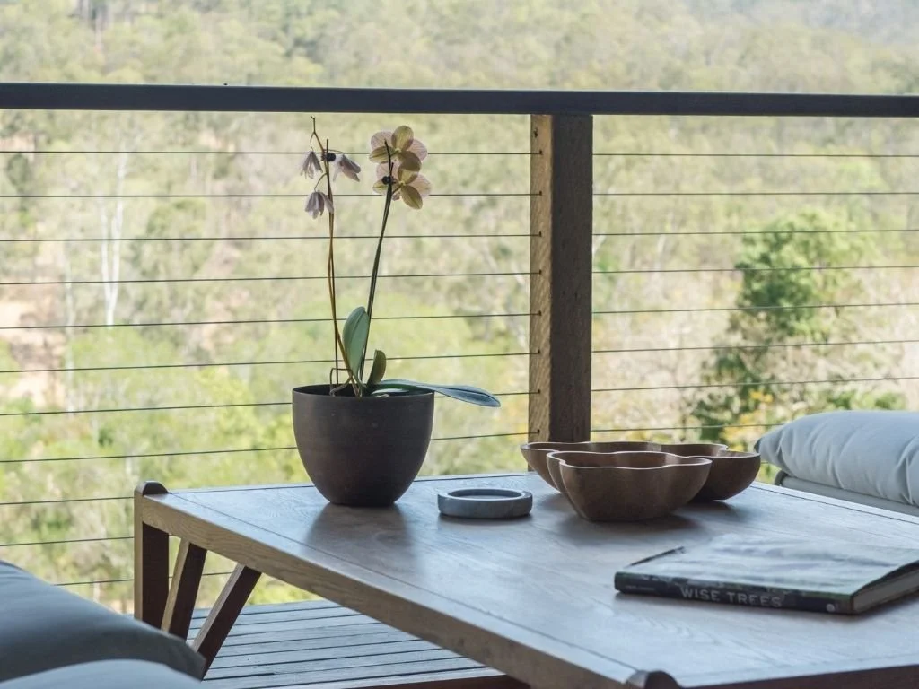 A wooden table with a potted orchid, a small dish, and four brown bowls on a balcony with a view of green trees and a wire railing.
