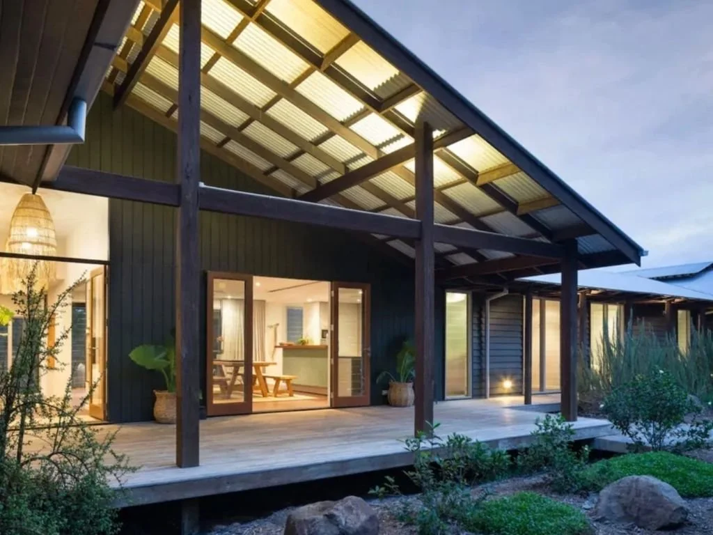 Modern house with a large wooden deck, dark exterior walls, and a sloped metal roof. Large sliding glass doors provide a view into the interior, which has a dining area with a table and benches. The house is surrounded by native plants and rocks.