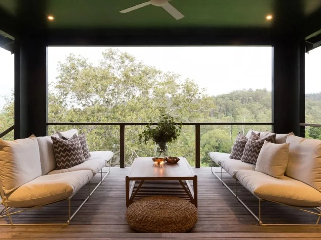 Outdoor balcony with two white sofas, a wooden coffee table with a potted plant, and a view of green trees and hills in the background.