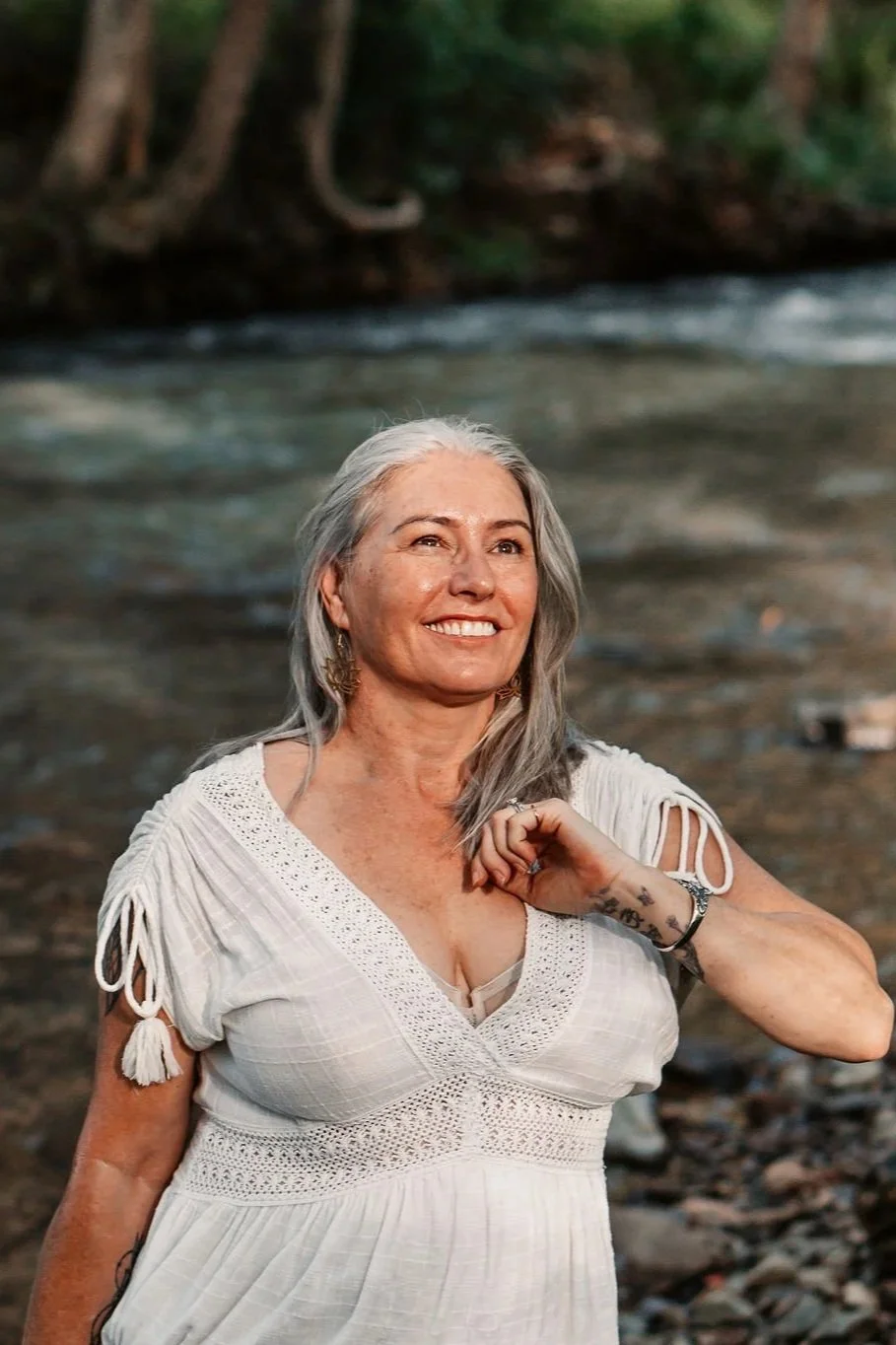 A woman with gray hair and a white dress standing near a river with a forest in the background.