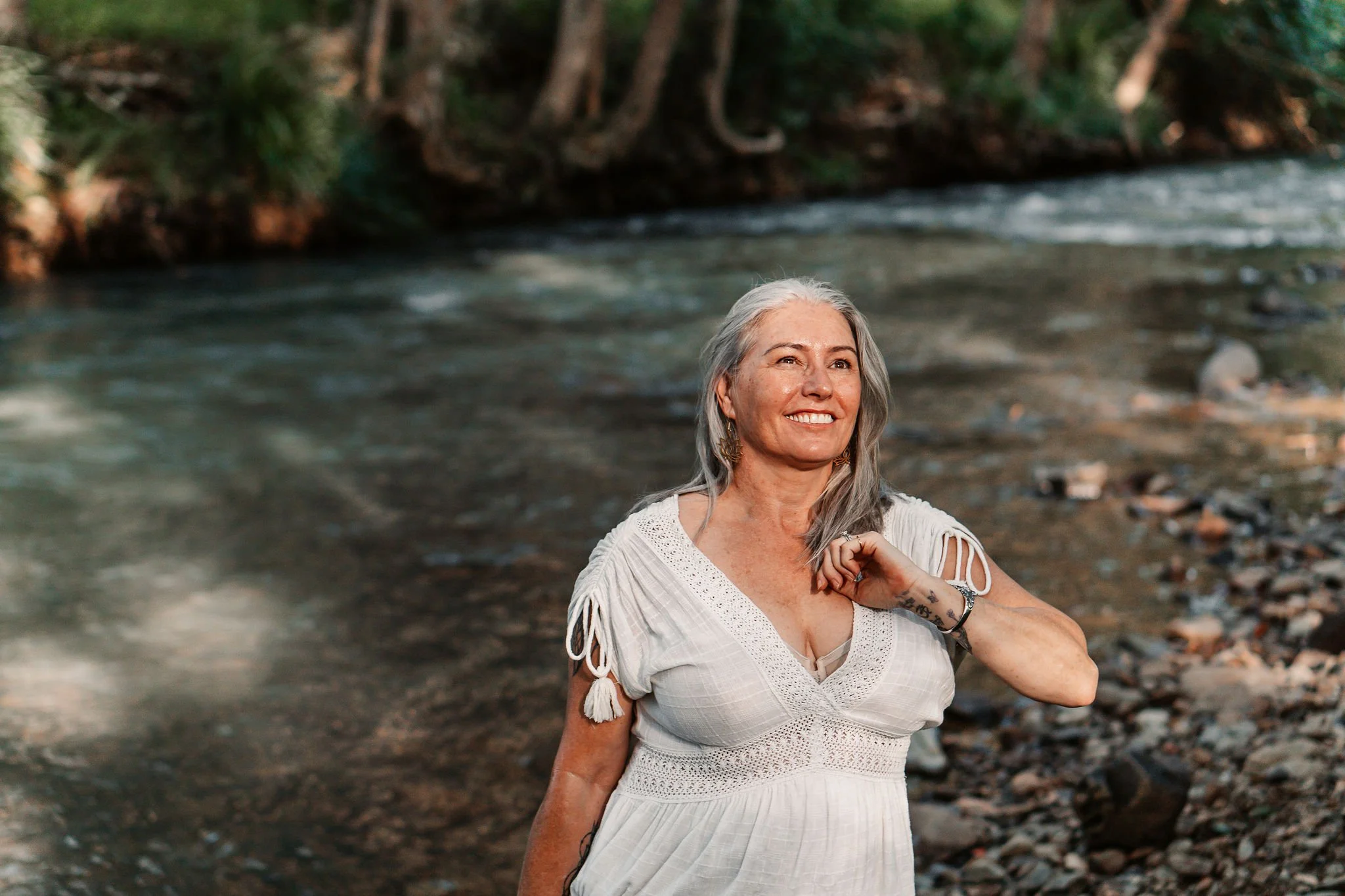 A smiling woman with gray hair standing near a riverbank with trees in the background.