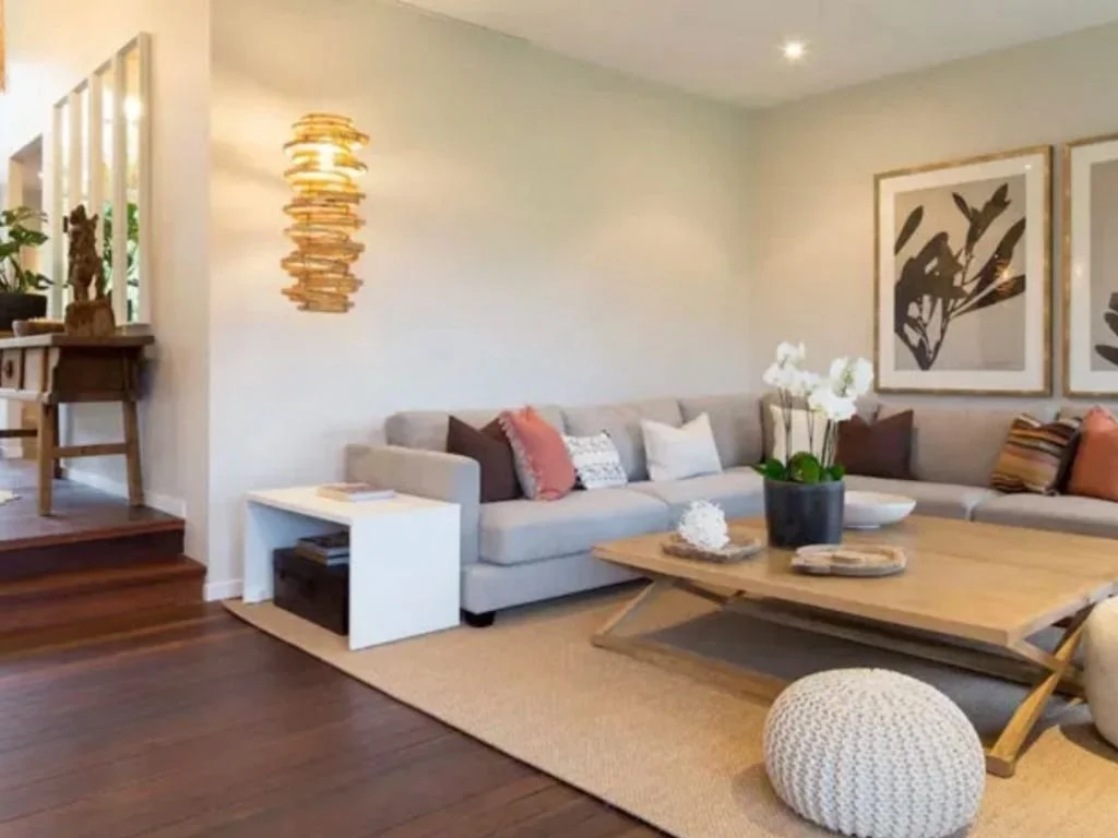 A living room with a cream sectional sofa, decorative pillows, a round knitted ottoman, a wooden coffee table with a potted orchid, framed artwork on the wall, and a wooden side table with a stack of books. There is a staircase partially visible to t