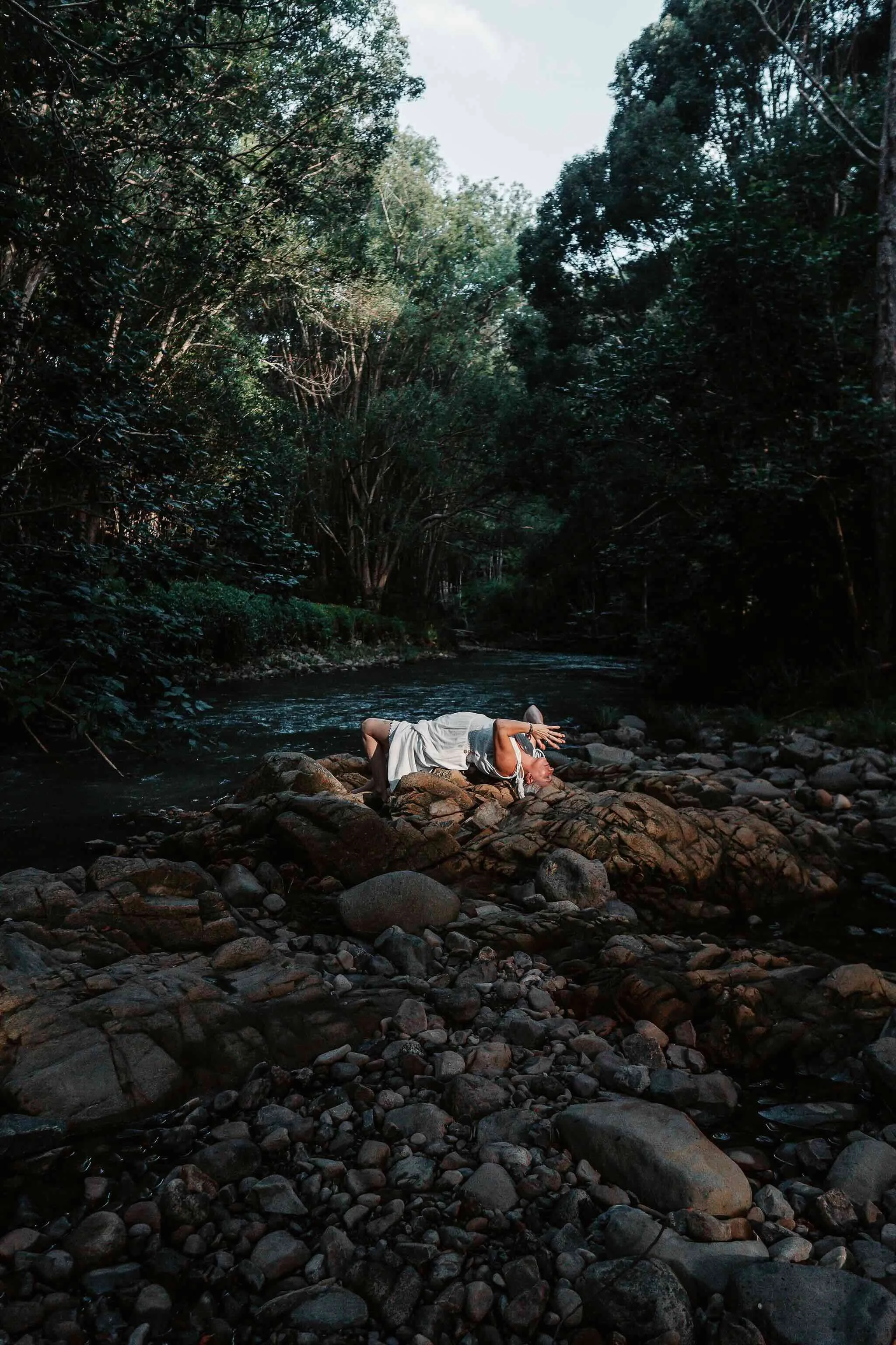 Person lying on rocks by a river in a forest, shielding their eyes from the sunlight with one hand.