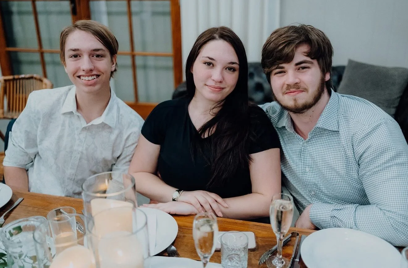 Three young adults sitting at a dinner table smiling for the camera, with plates, glasses, and a lit candle visible on the table in a cozy indoor setting.