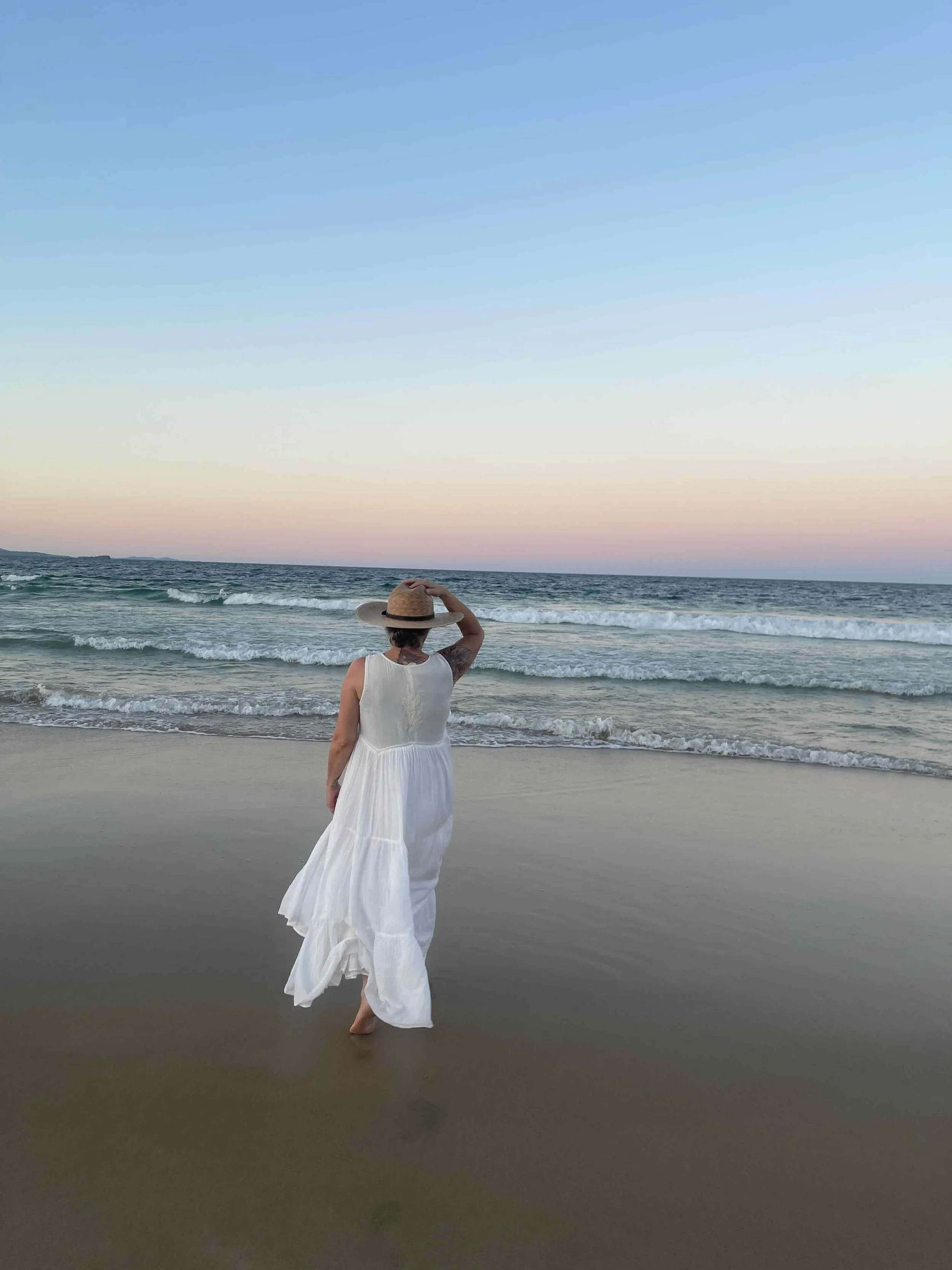 Woman in a white dress and straw hat walking on the beach near the ocean during sunset.