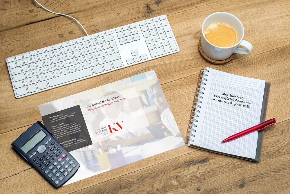 Desk with a white keyboard, a cup of coffee, a calculator, a spiral notebook with a handwritten note, and a school asbestos management plan
