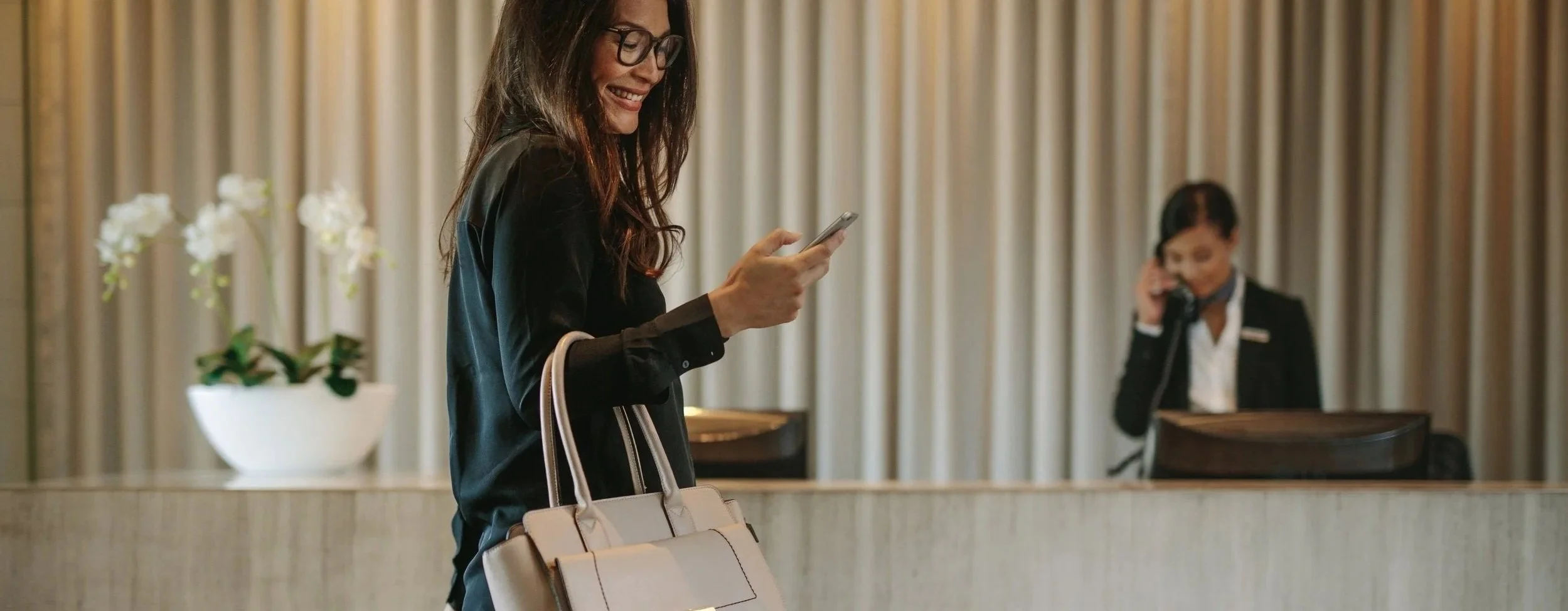 A woman with glasses looking at her phone and smiling, standing at a hotel reception desk with a bellhop in uniform in the background, talking on a phone.