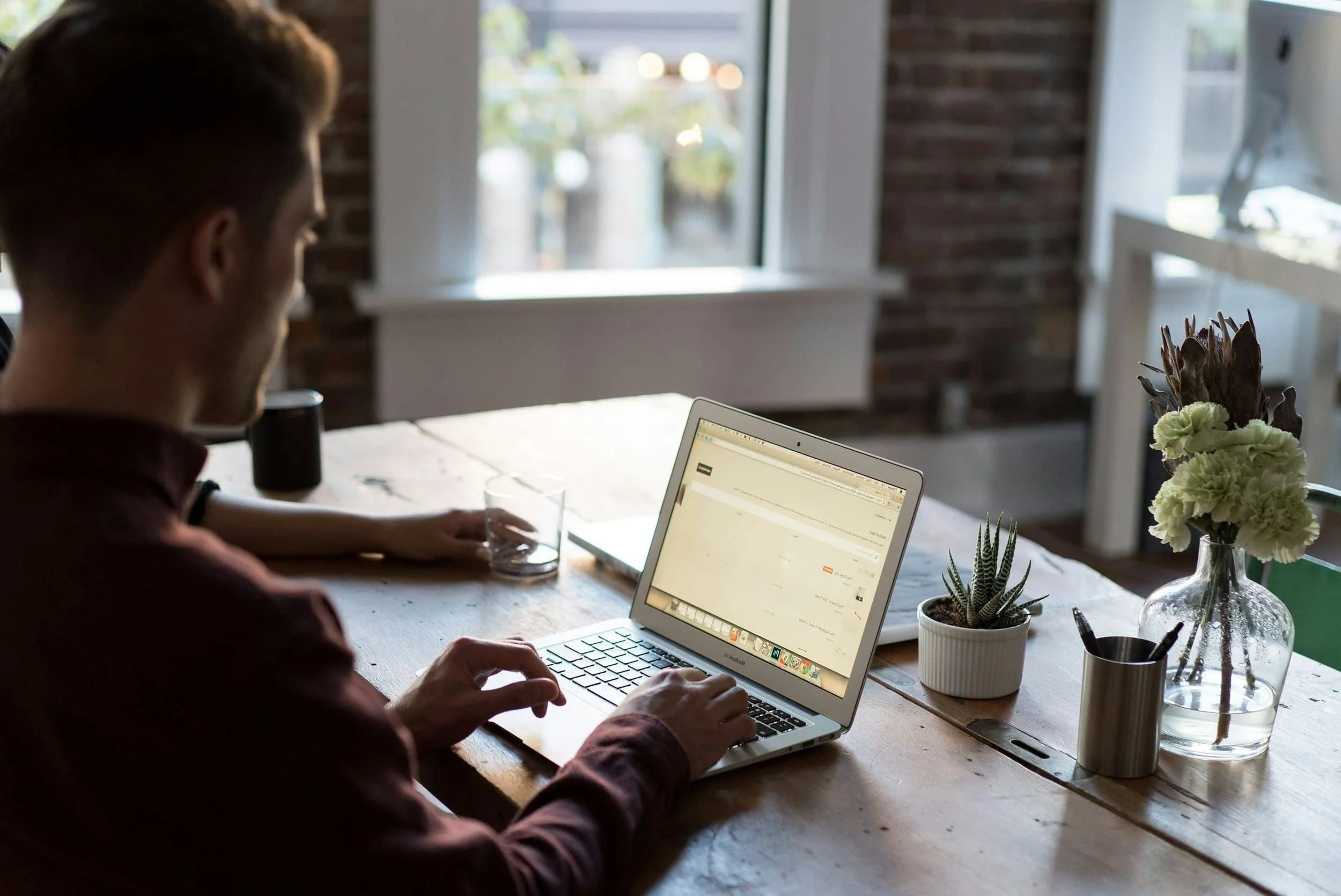 Person working on a laptop at a wooden table inside a room with large window, plants on the table, and a view outside.