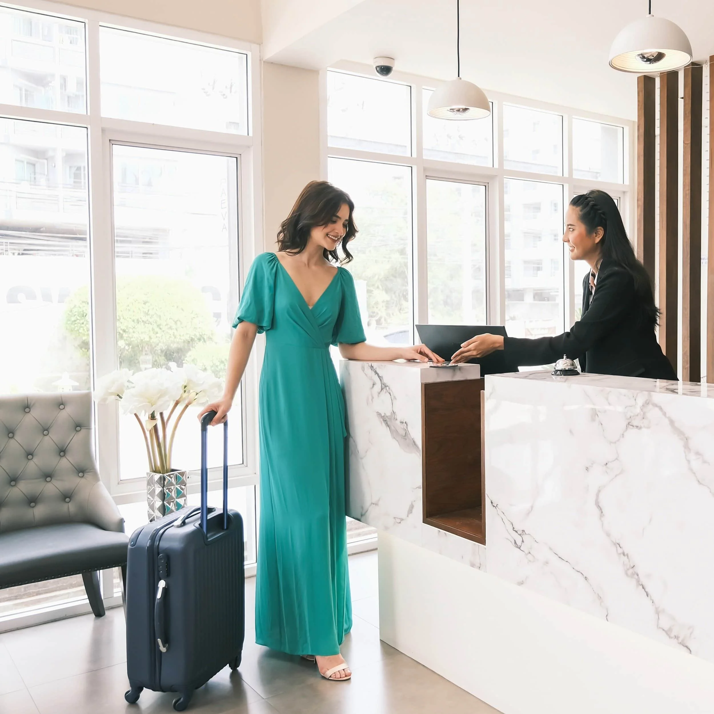 A woman in a turquoise dress with a rolling suitcase at a hotel reception, checking in with the receptionist.