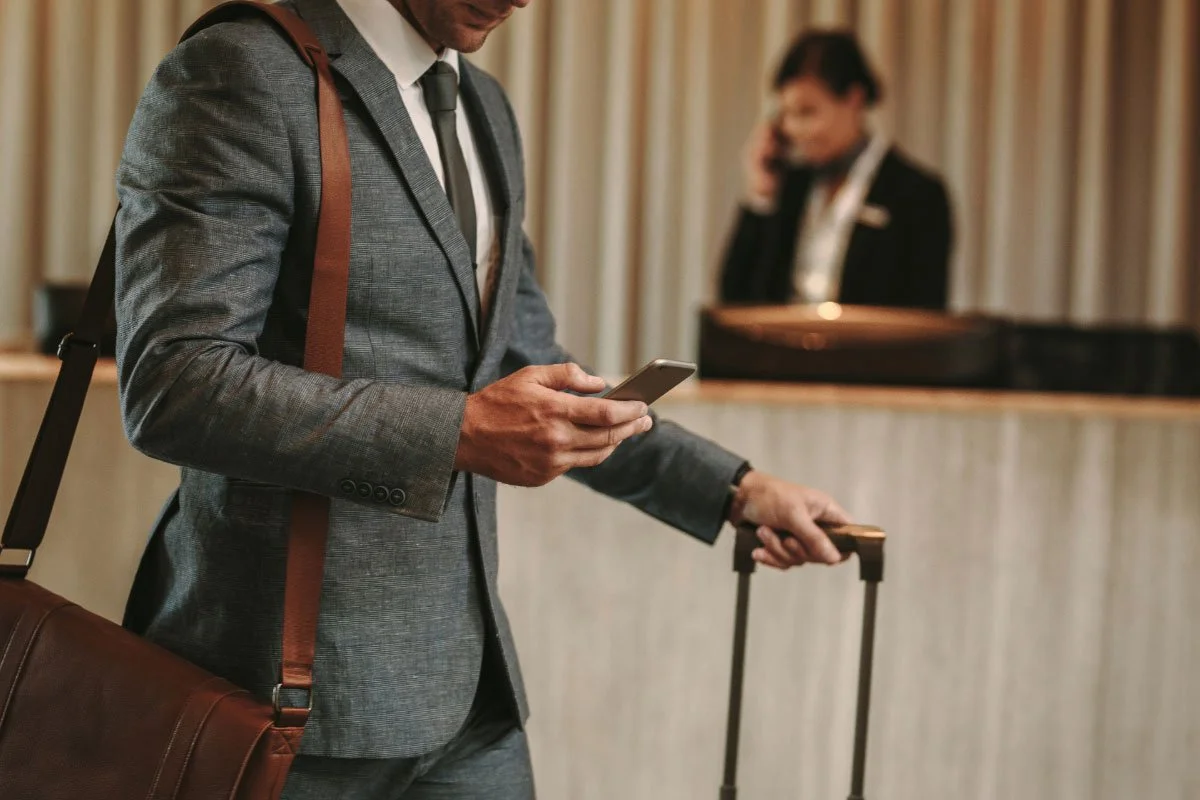 Man in a gray suit with a brown shoulder bag using a smartphone at a hotel reception desk, with a woman on the phone in the background.