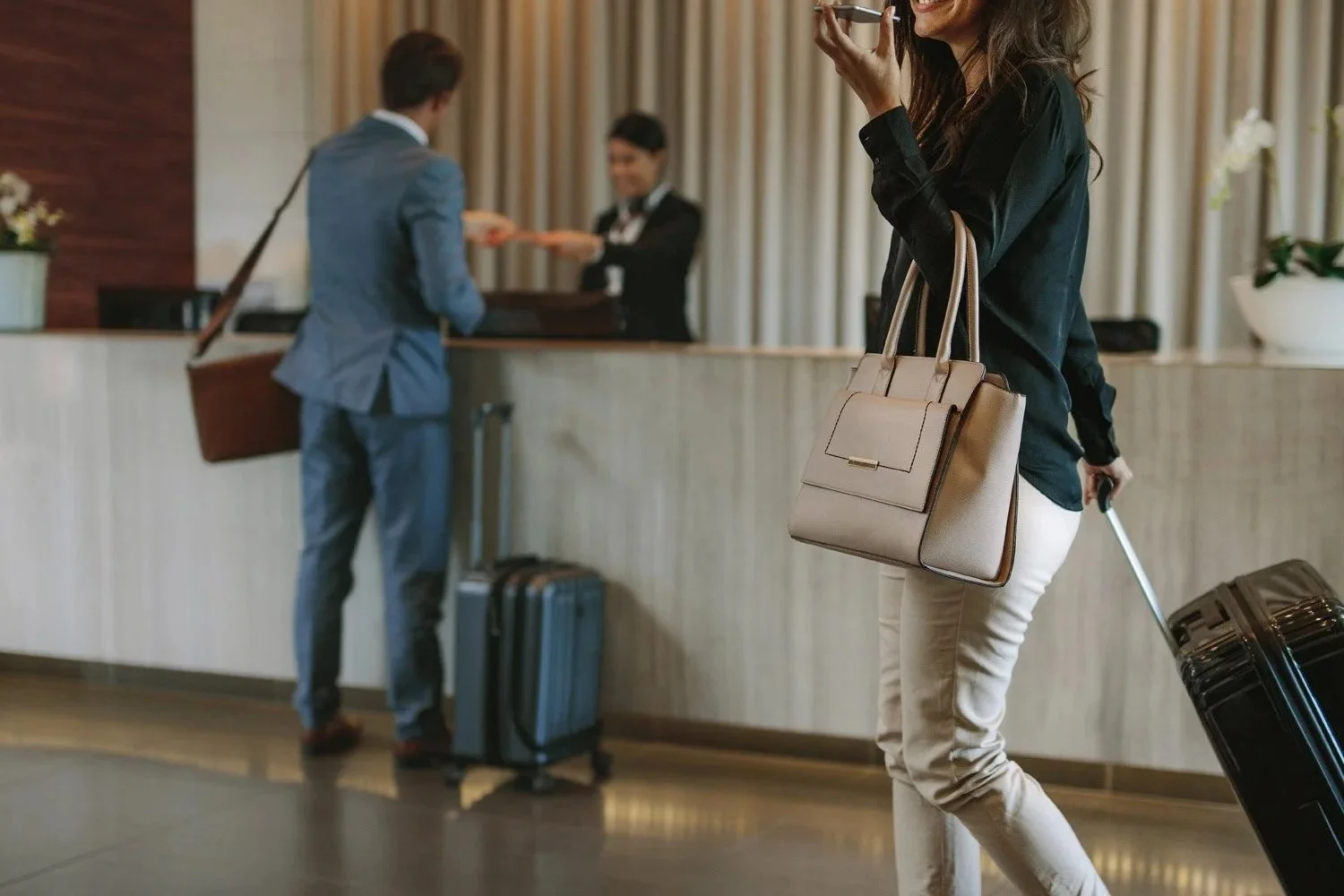 A woman at a hotel check-in desk pulling a black suitcase, carrying a beige handbag and holding a phone, with a man in a suit and an employee behind the counter in the background.
