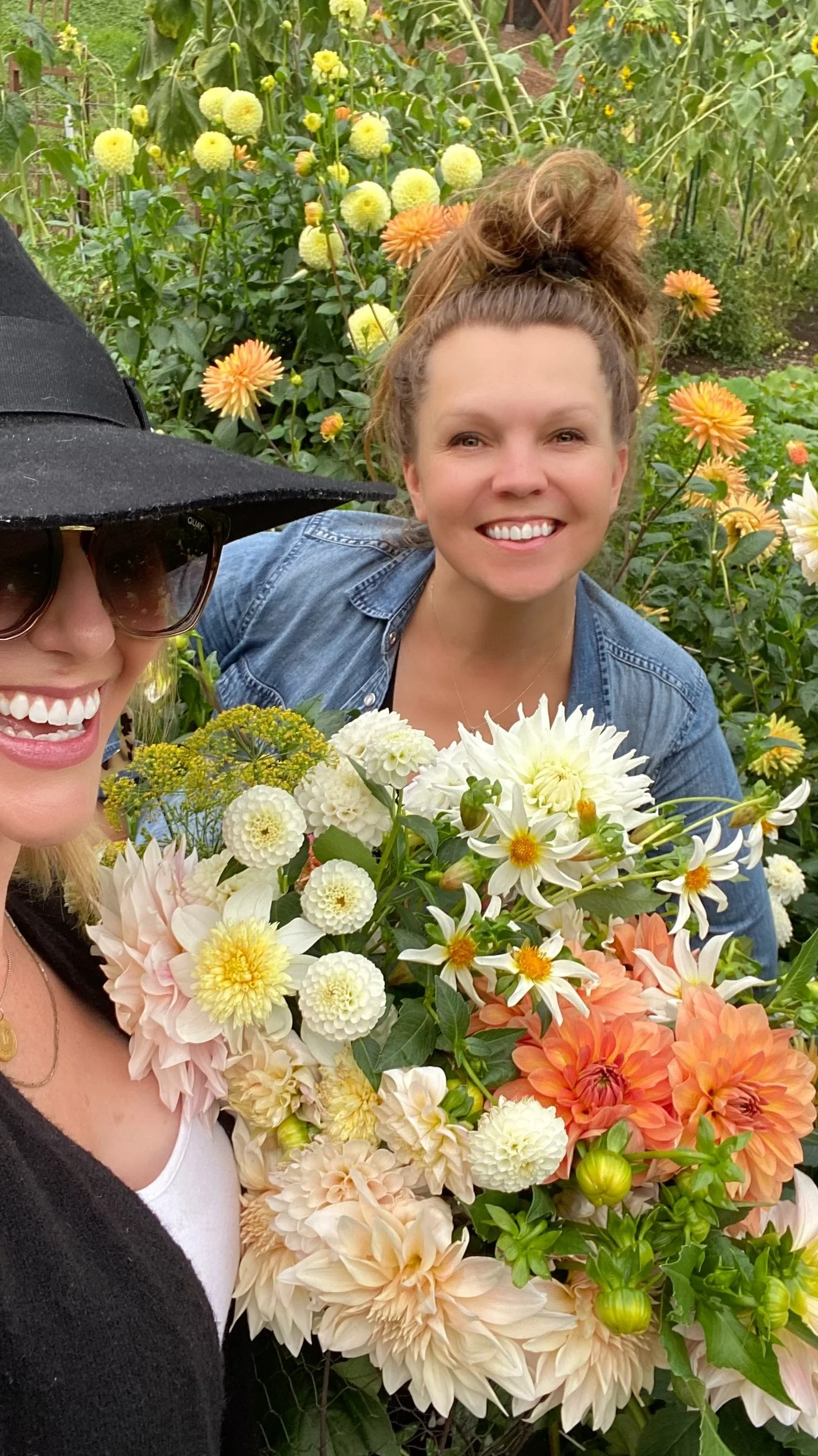 Two women smiling in a garden with a large bouquet of colorful flowers, including dahlias and daisies; lush greenery in the background.
