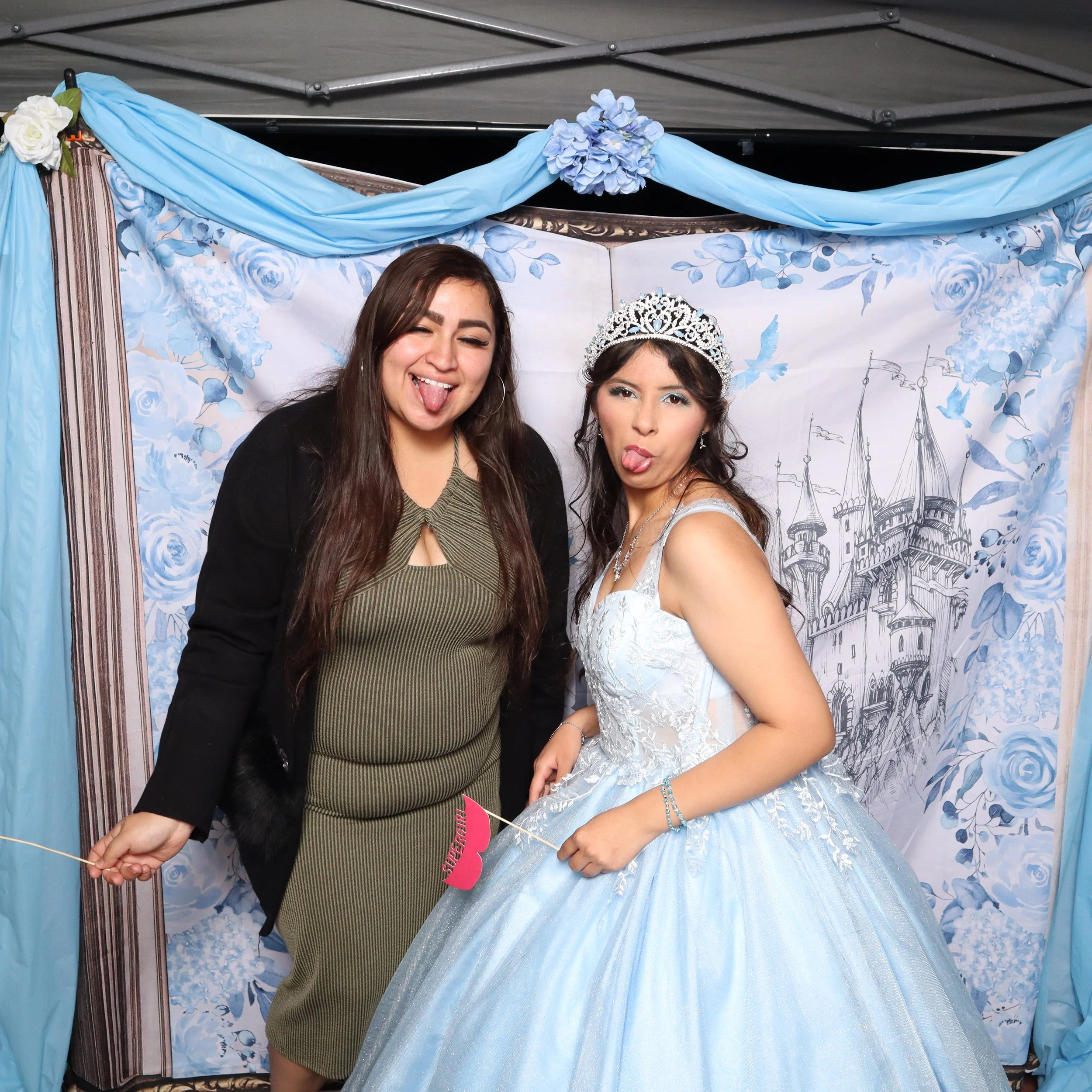 Two women posing playfully in front of a fairy tale-themed backdrop, one dressed as a princess with a tiara and gown, and the other dressed casually, making funny faces.