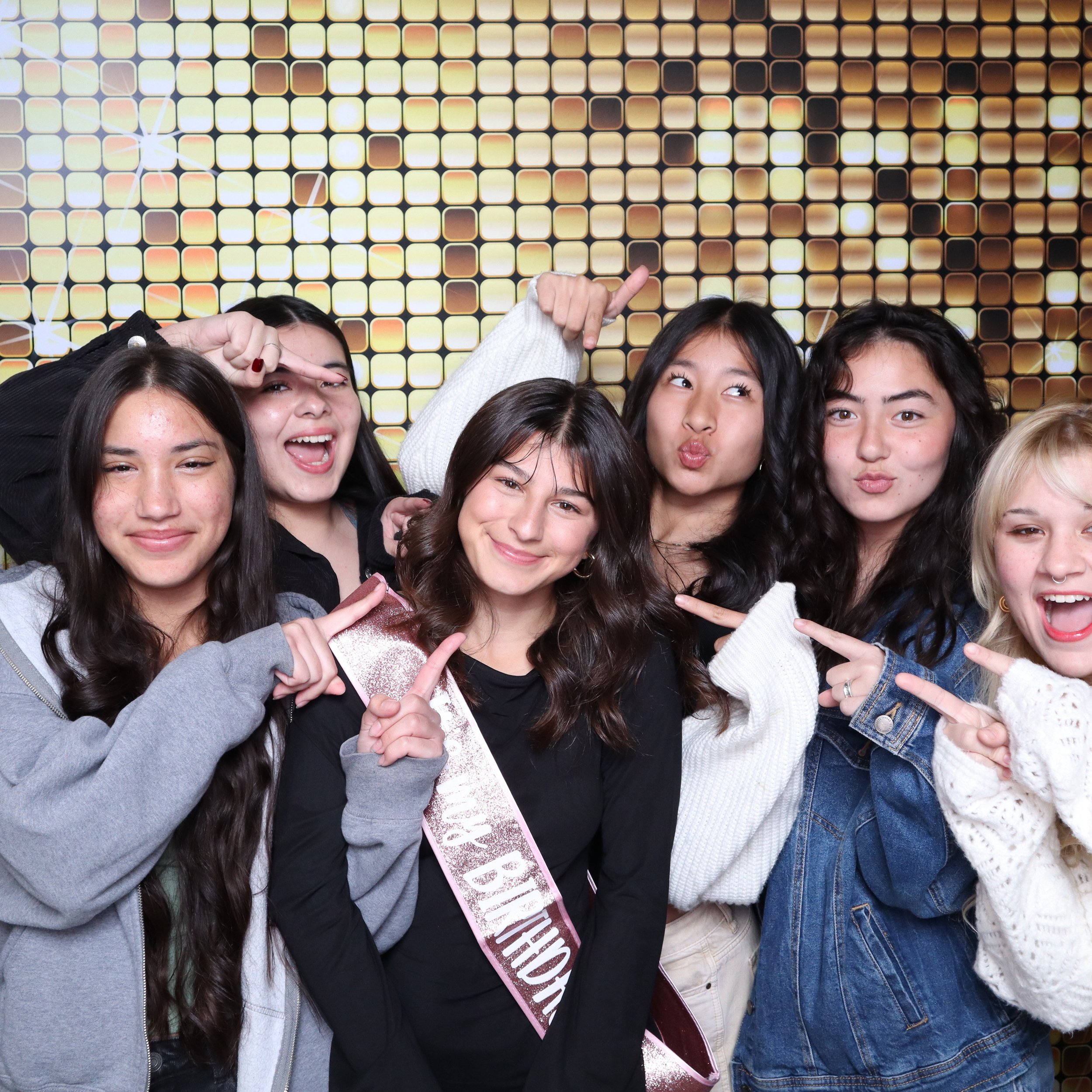 Group of six young women celebrating at a party with a golden, glittering backdrop. One woman is wearing a sash that reads "PROM BIRTHDAY," and others are pointing or making gestures while smiling and making playful faces.