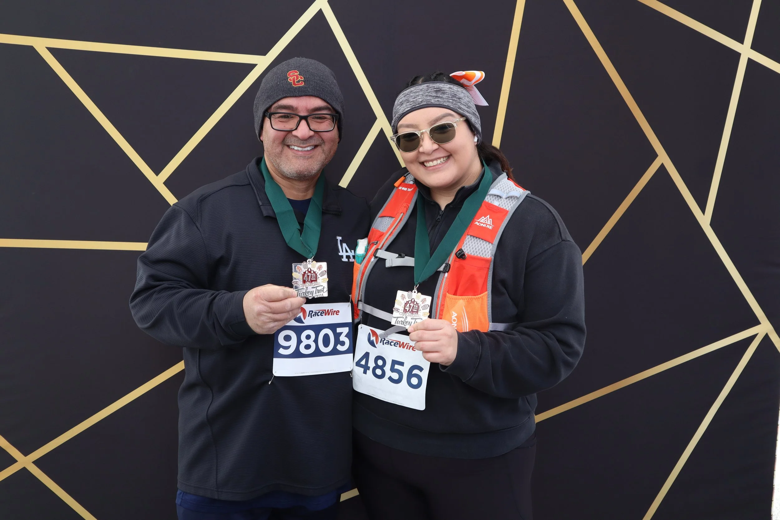 Two smiling marathon runners holding up medals, standing in front of a black backdrop with gold geometric lines, wearing race bibs and athletic clothing.