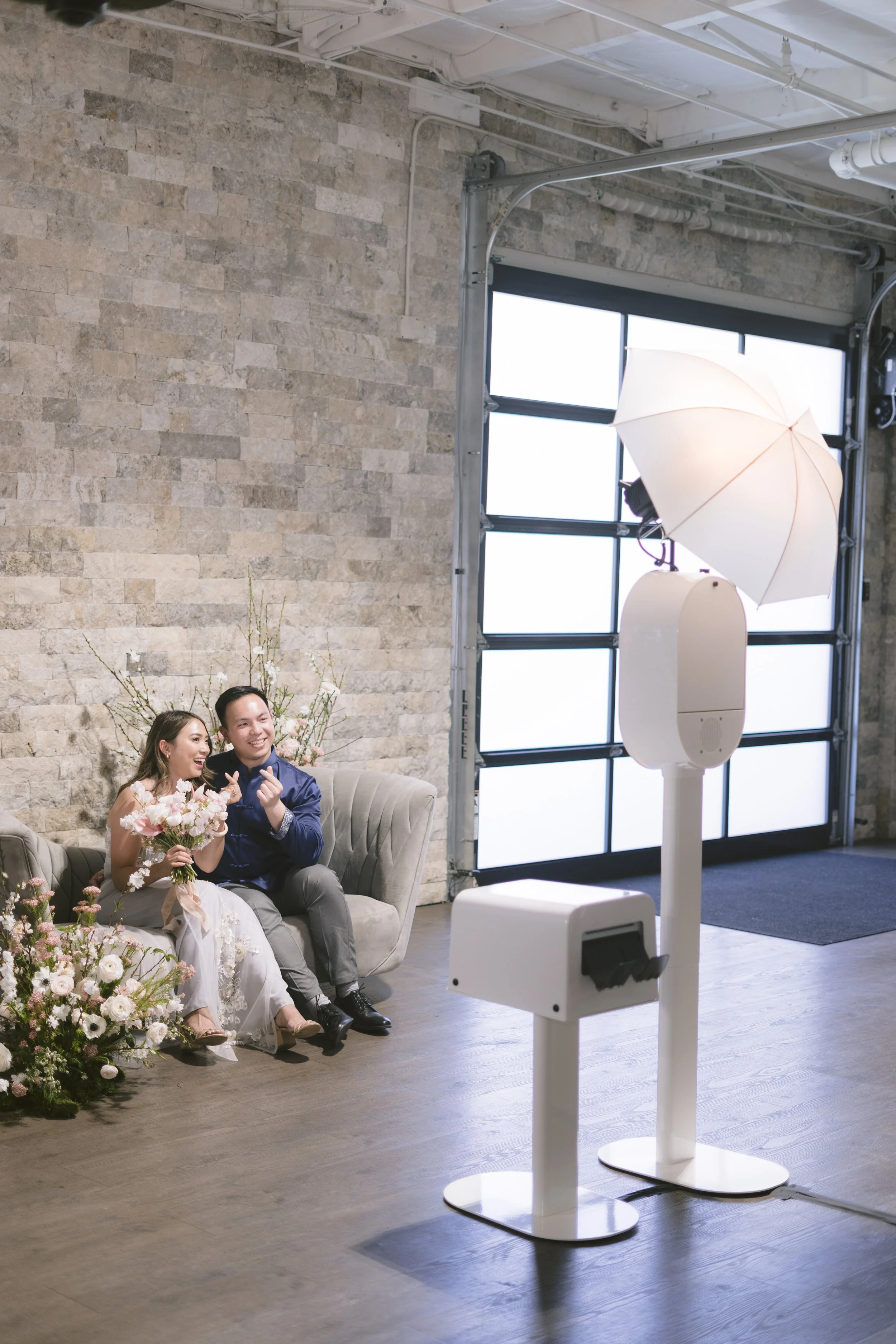 A bride and groom sitting on a gray couch, smiling and holding flowers, in a photo studio with a photo booth setup, including a large hairdryer-shaped light, an umbrella, and a backdrop, with an industrial-style brick wall and large garage door in the background.
