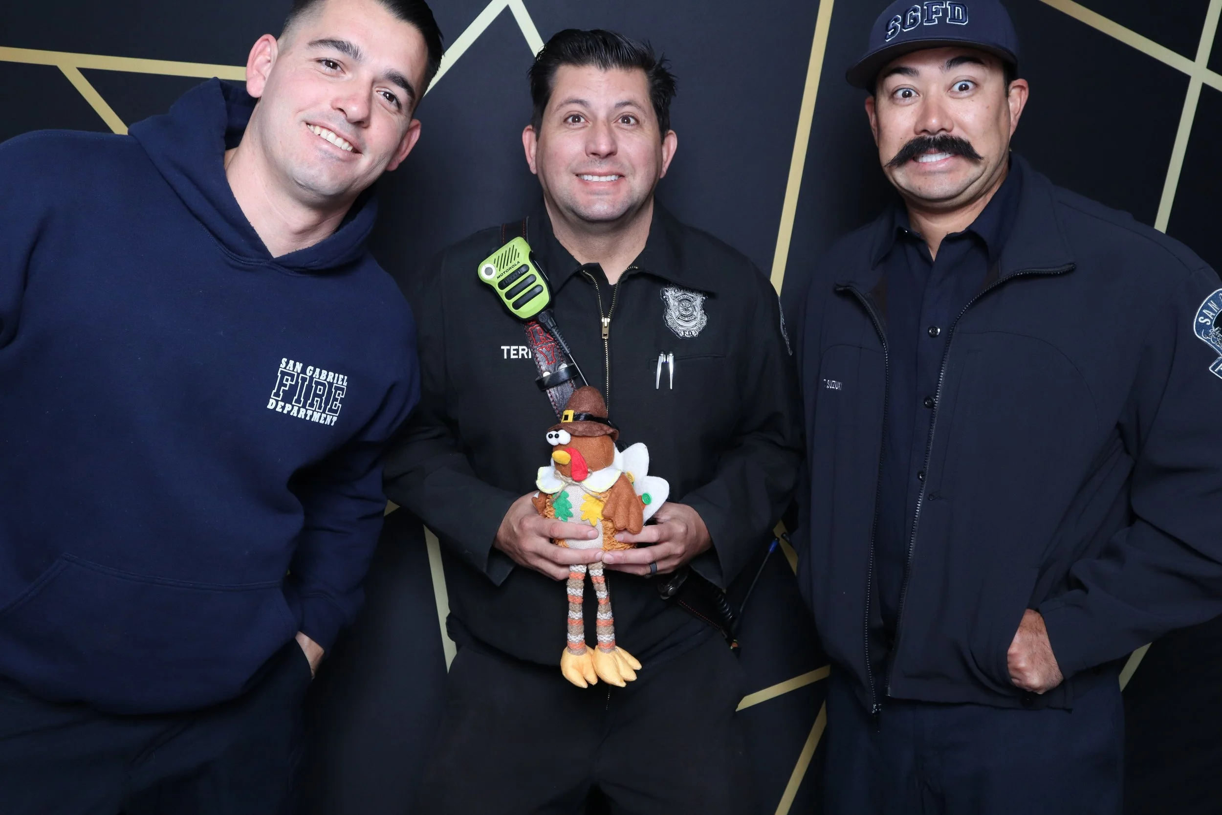 Three men in fire and police uniforms standing together, one holding a turkey plush toy.