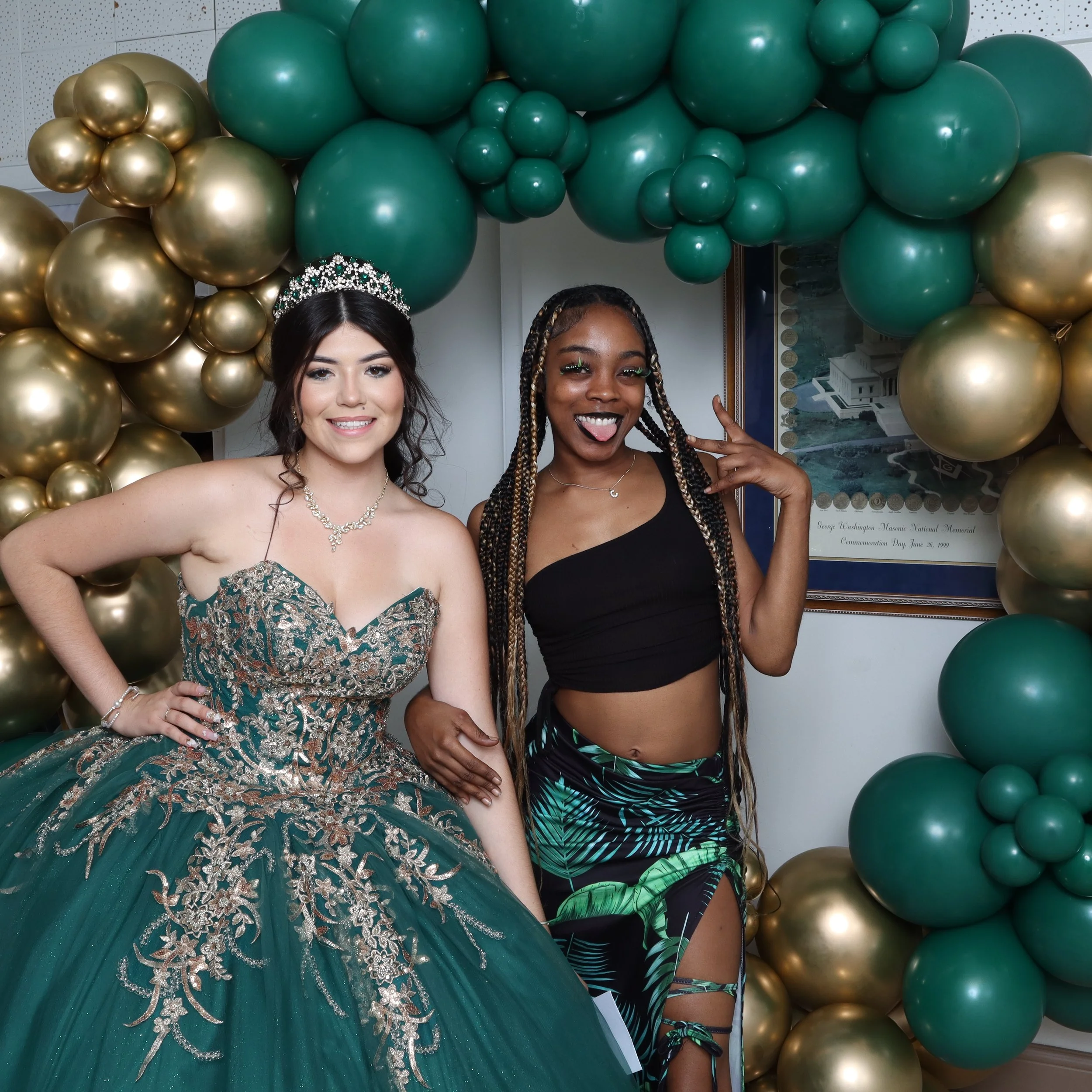 Two young women posing together at a celebration, surrounded by green and gold balloons. One woman is dressed in a green, gold-embellished gown with a tiara, and the other is in a black crop top with a tropical print skirt, making a peace sign and st