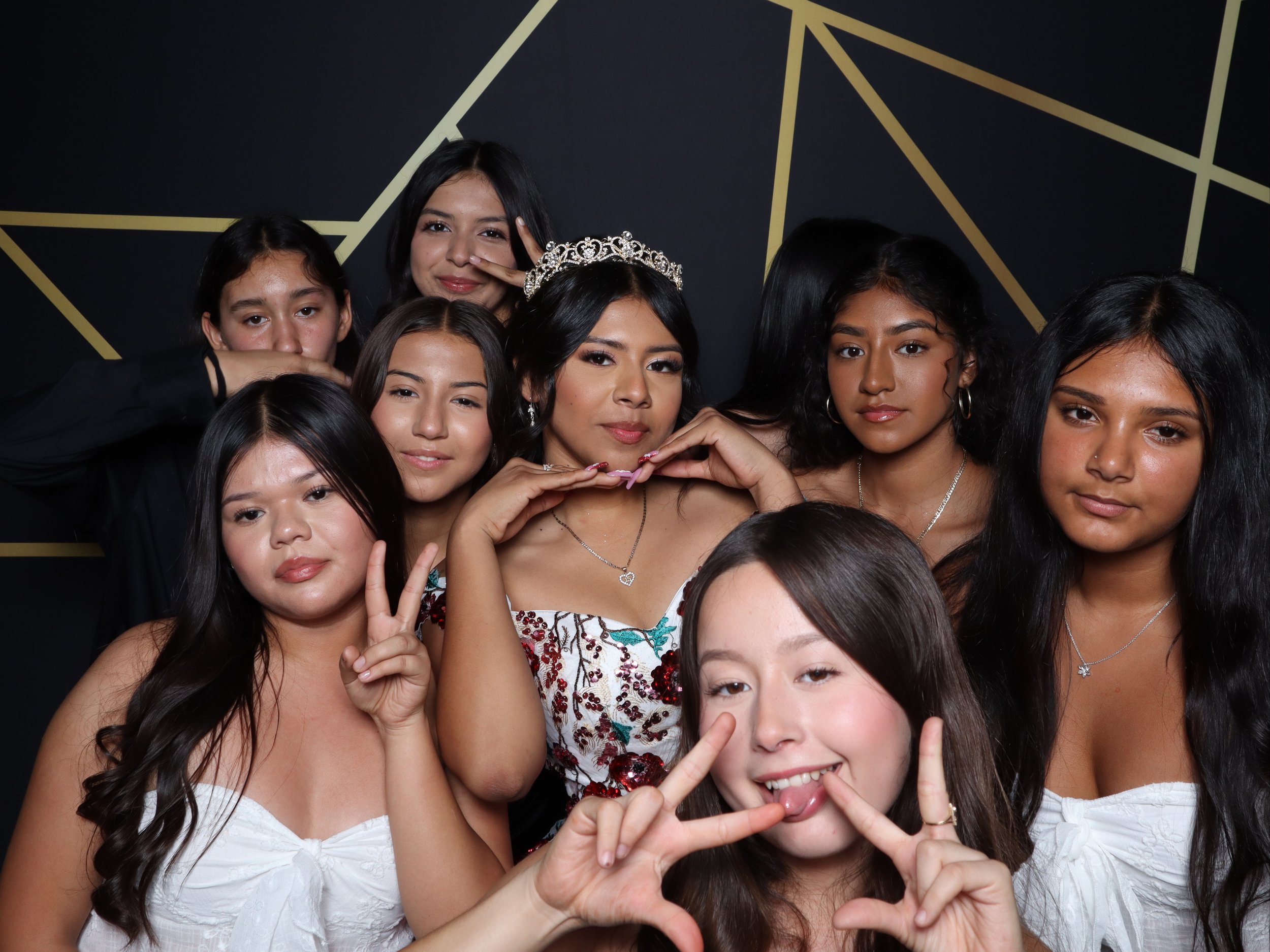 Group of nine young women, one wearing a tiara, posing for a photo in front of a black and gold geometric patterned background. They are making peace signs and cute gestures with their hands.
