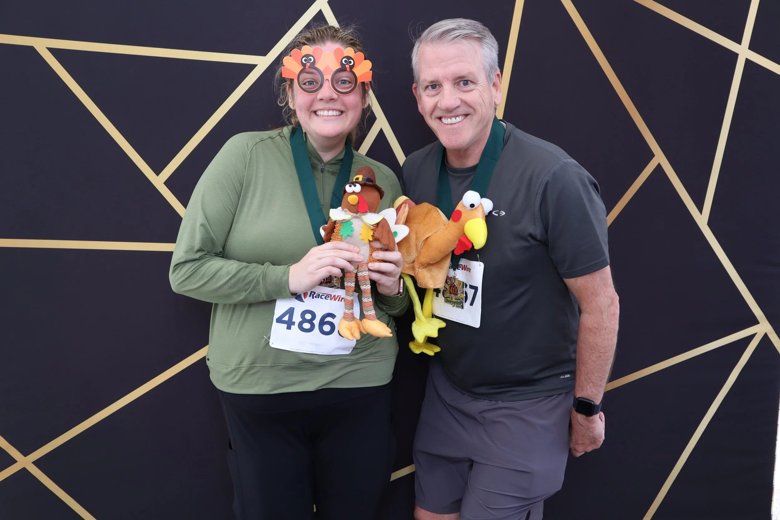 Smiling woman and man wearing race bibs pose together at a race event, holding plush turkey and Thanksgiving-themed stuffed toys, with a black and gold geometric patterned backdrop.