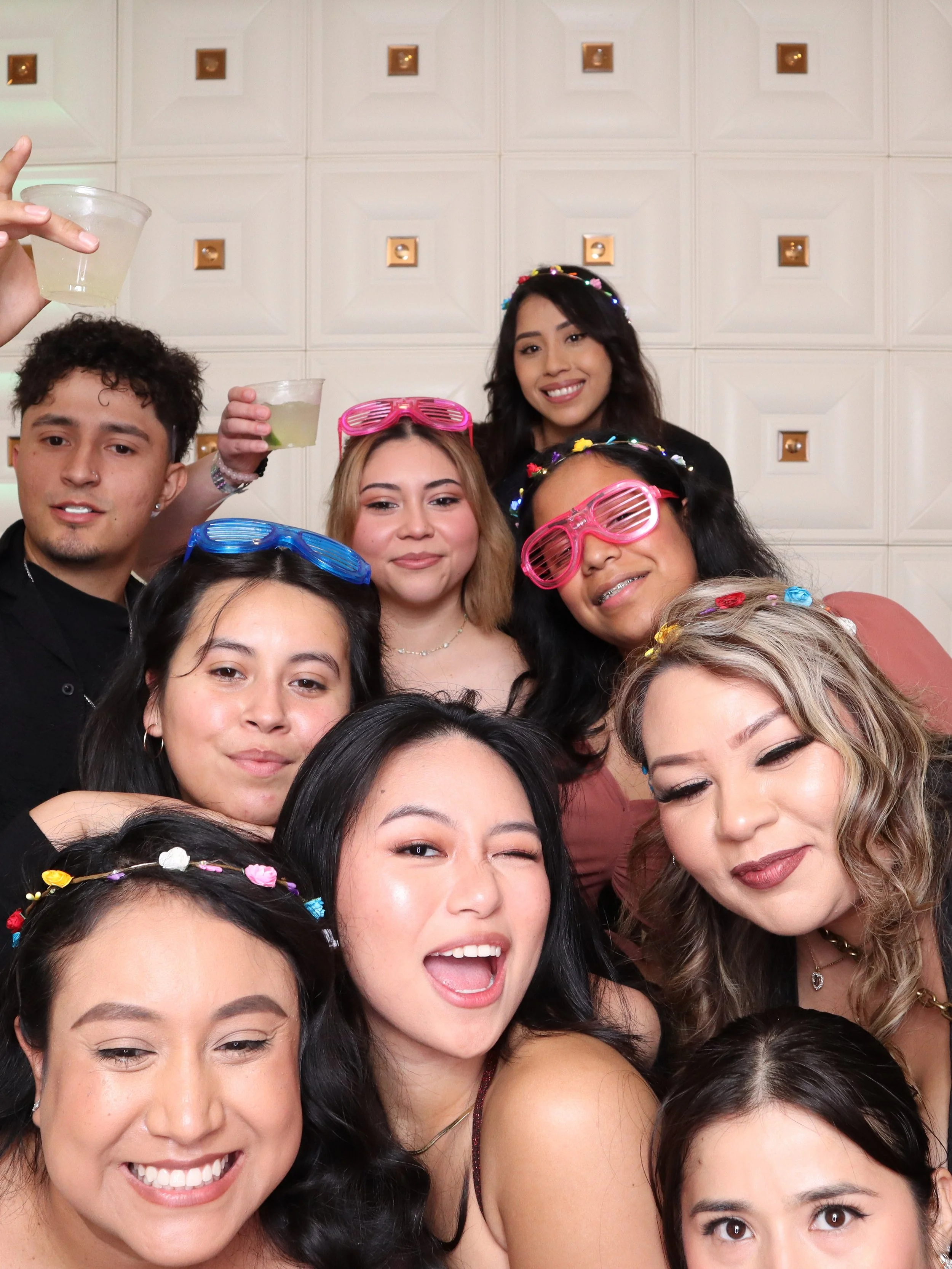 Group of young adults celebrating at a party, some wearing colorful glasses and floral headbands, posing for a photo with drinks in hand.