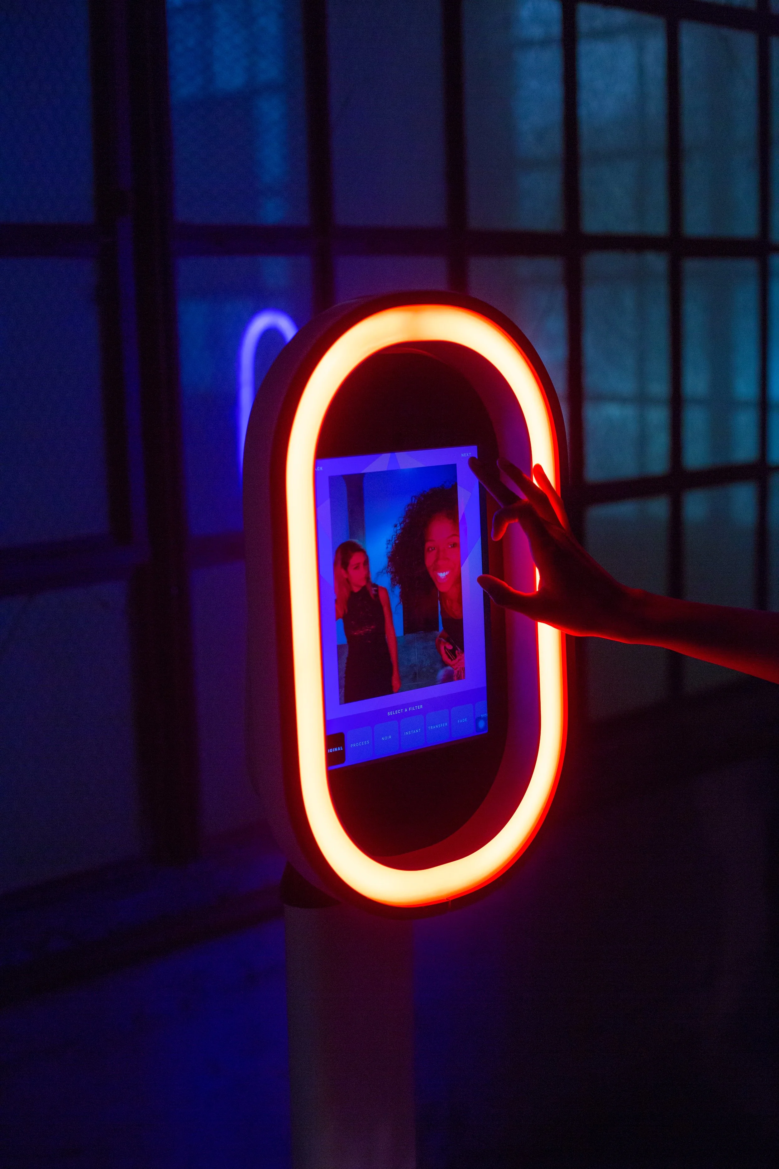 A person is using a photo booth with a glowing red and orange oval frame and a digital touchscreen display, taking a picture of themselves and a woman with curly hair.