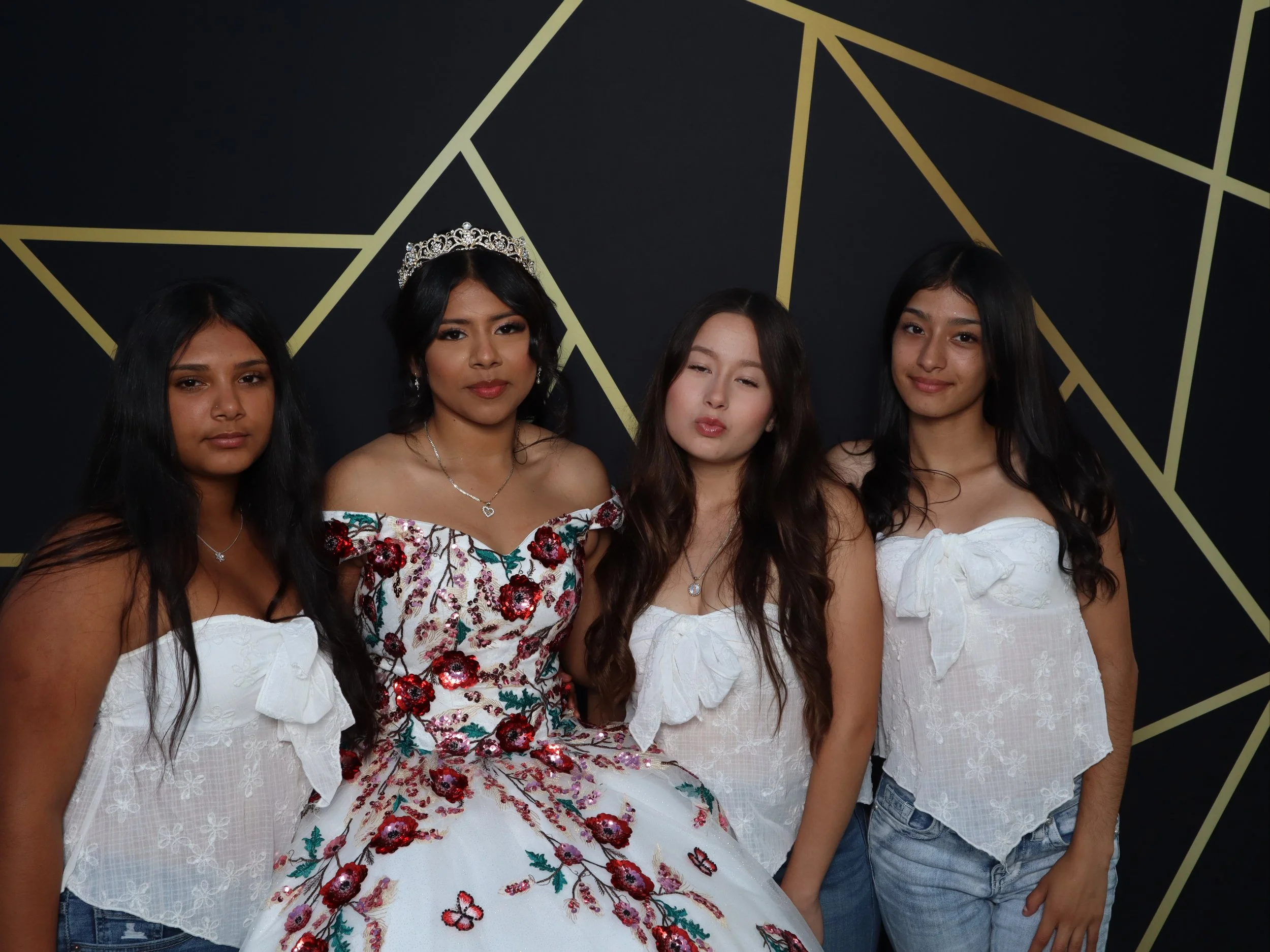 Four young women standing together in front of a black wall with gold geometric star pattern. The second woman from the left is dressed in a fancy floral ball gown with a tiara, while the others wear casual white tops and jeans.