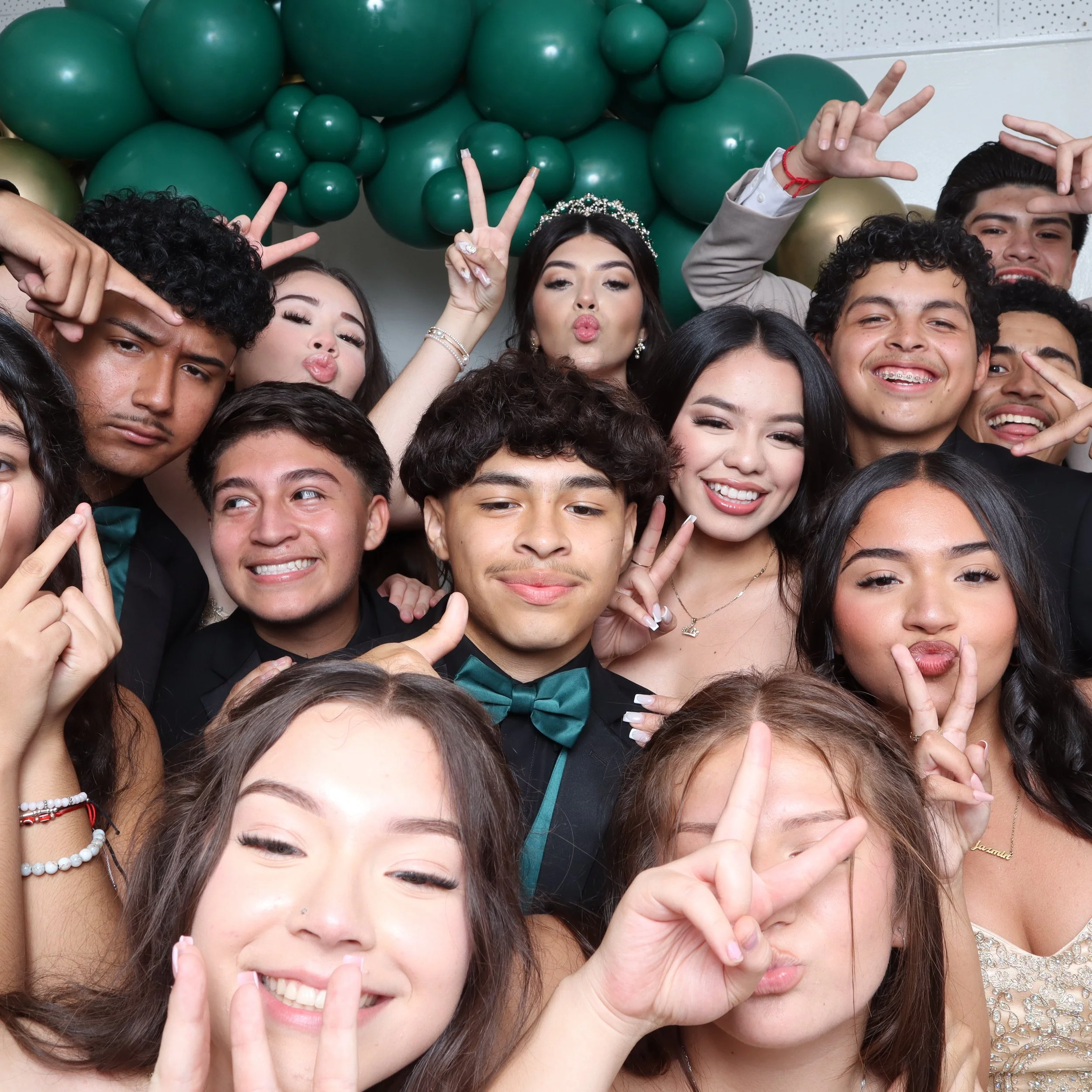 A group of young people at a celebration, with a woman in the center wearing a tiara, all making peace signs or playful gestures in front of green and gold balloons.