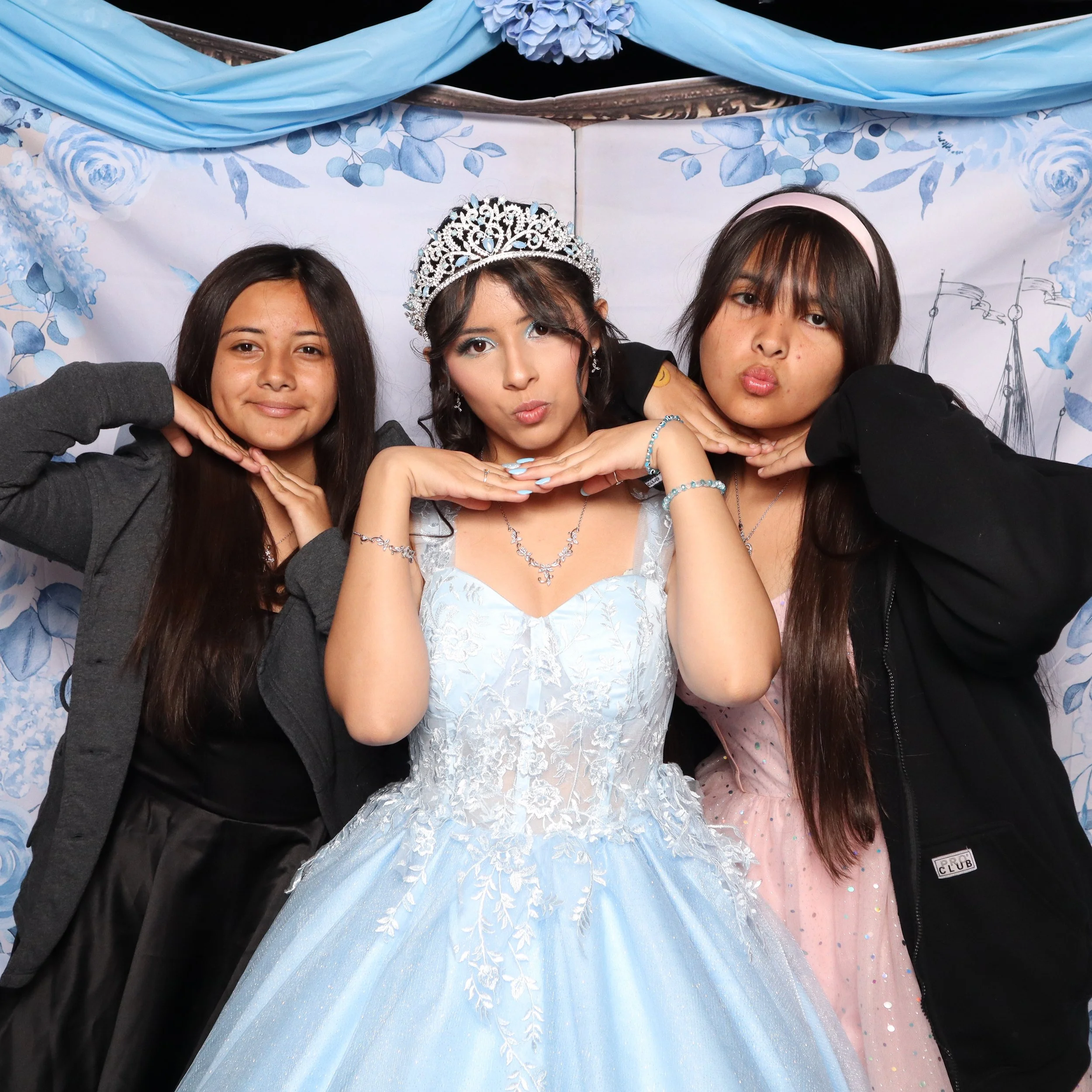 Three young women posing together, with the girl in the middle dressed as a princess or quinceañera, wearing a tiara and a blue gown, while the other two girls are in casual clothes.