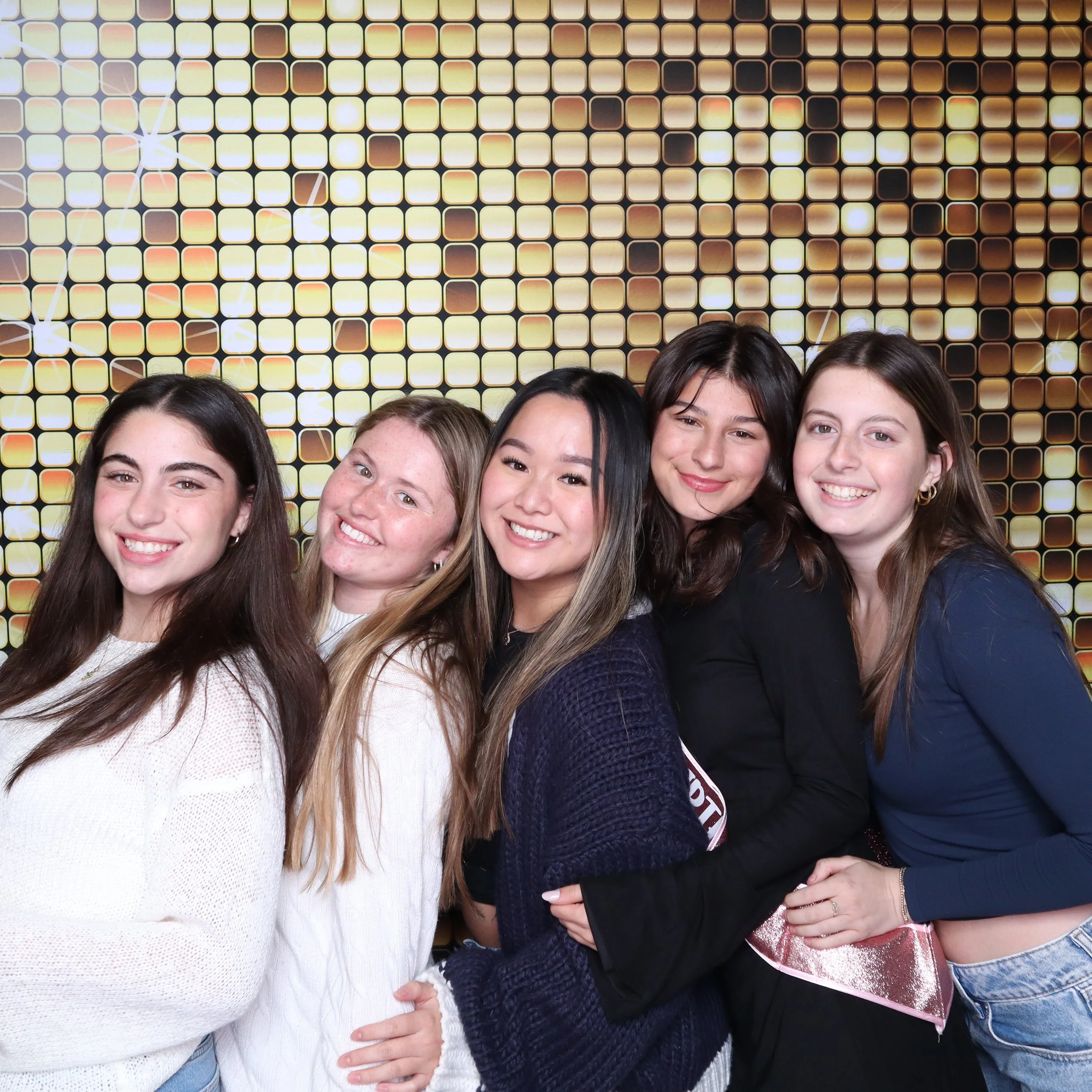 Group of five young women smiling and posing together in front of a gold and black reflective backdrop.