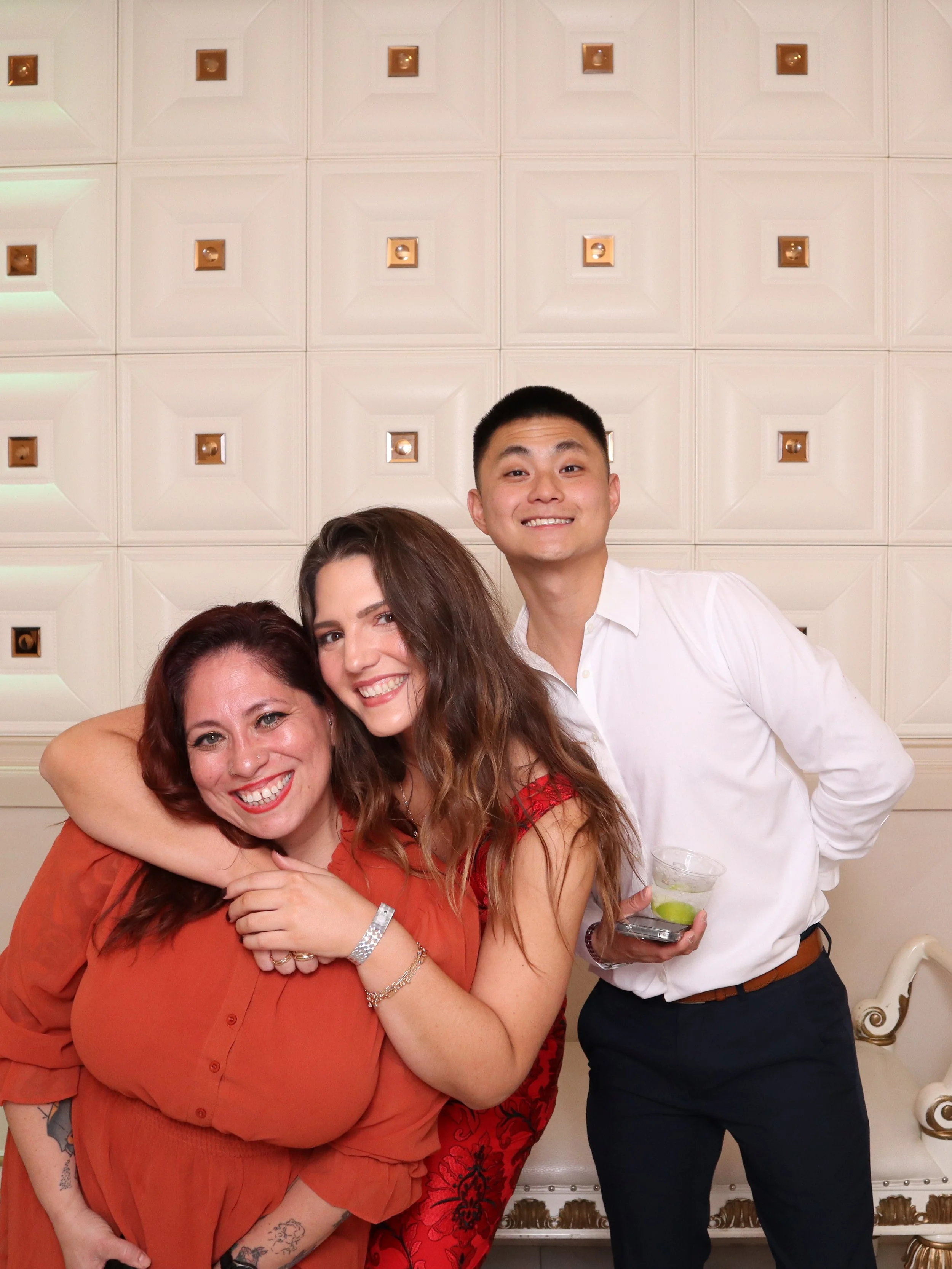 Three smiling people, two women and one man, posing together indoors in front of a patterned wall. The women are hugging, and the man is holding a drink.