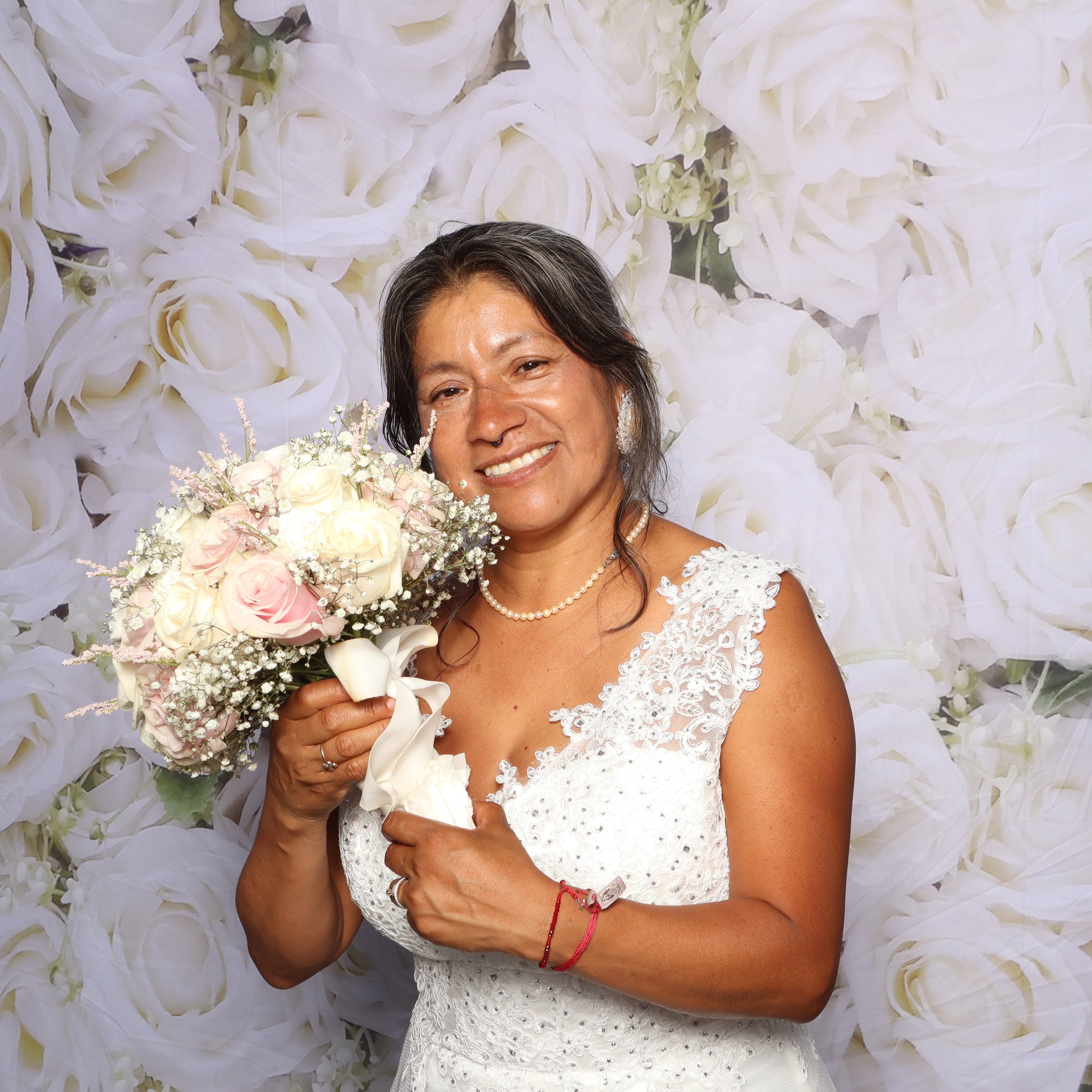 A woman in a white lace dress holding a bouquet of white and pink roses, smiling at the camera, with a floral backdrop.