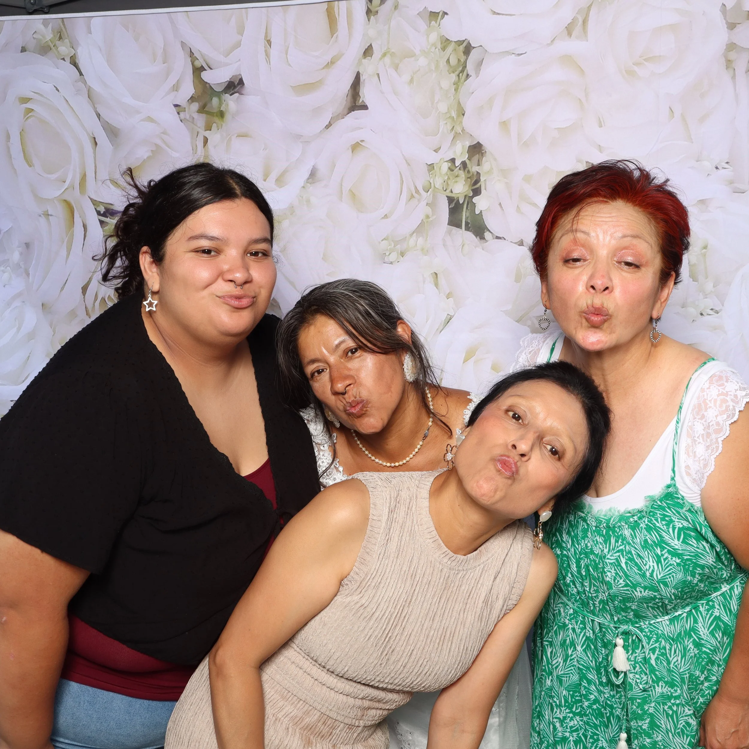 Group of five women making kissy faces in front of a white floral backdrop, dressed casually, wearing earrings and necklaces.