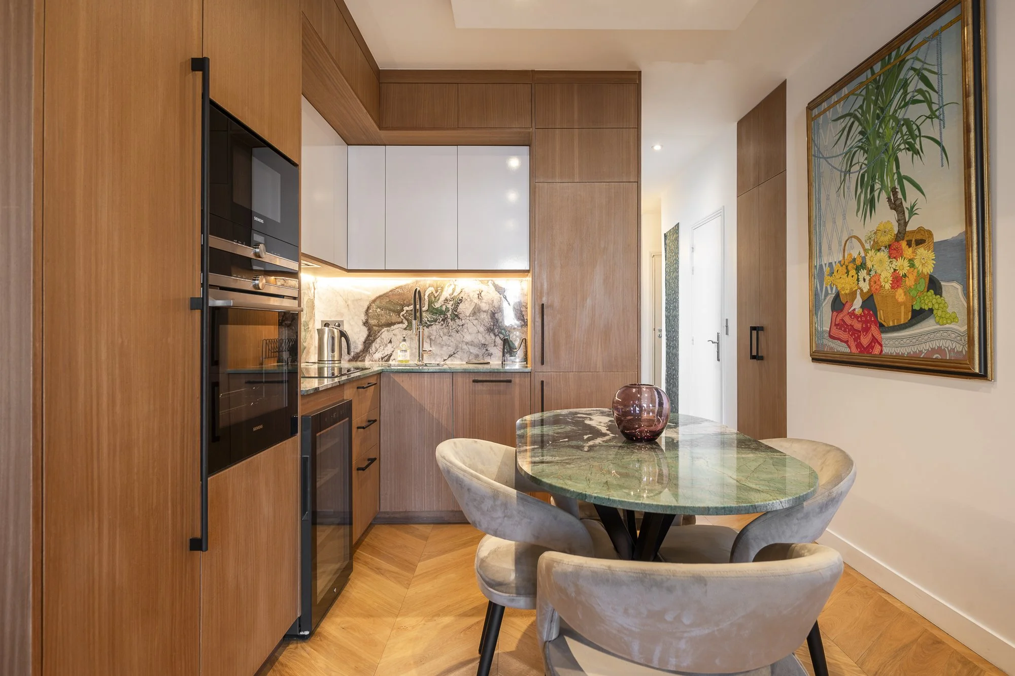 Modern kitchen with wooden cabinets, a marble backsplash, and a small round dining table with beige chairs. There's a colorful painting on the wall.