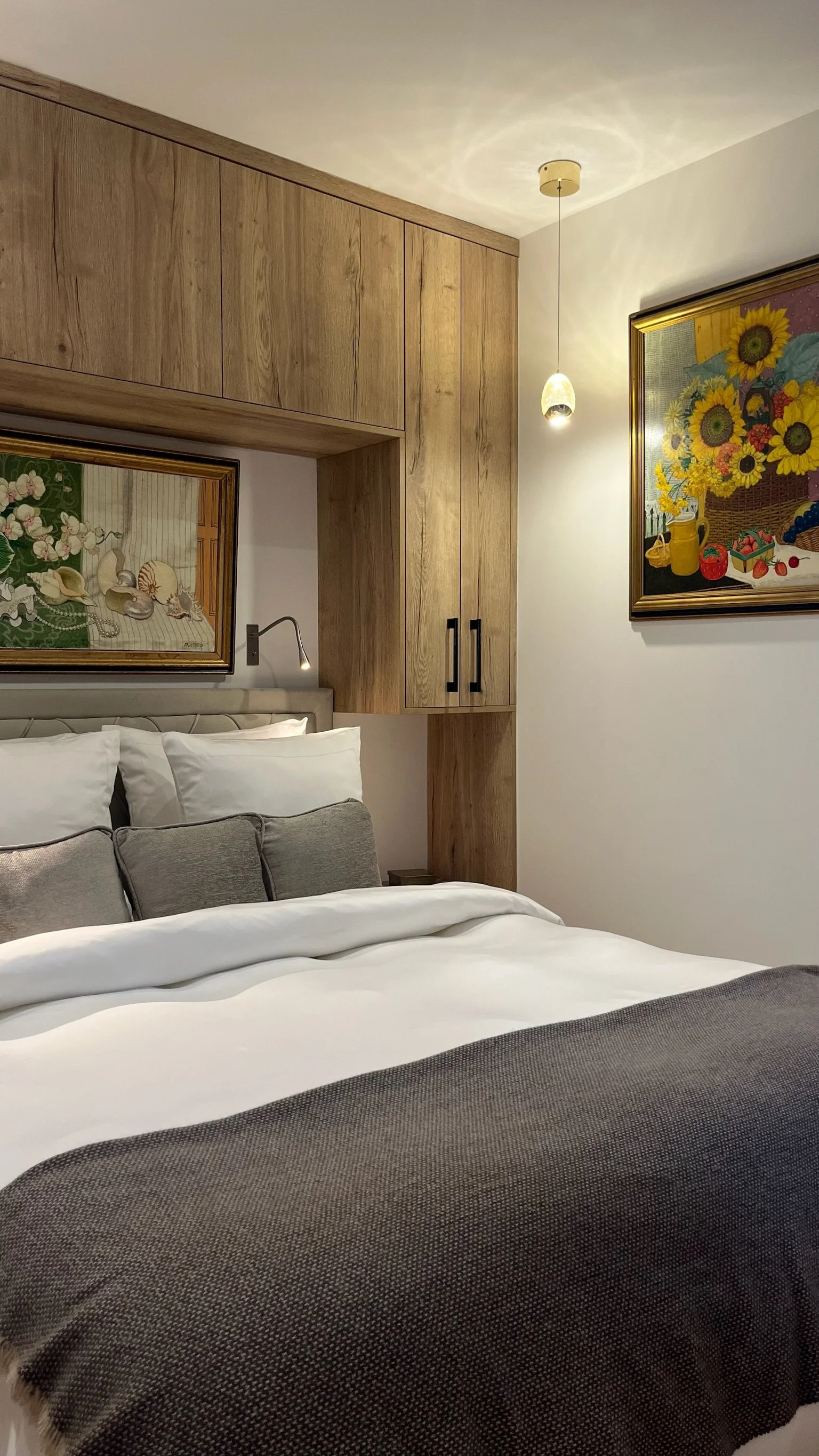 A modern bedroom with wood paneling and a cozy bed. The room features a wooden wardrobe with glass doors, white linens, a gray throw, and multiple pillows. A colorful painting of sunflowers hangs on the wall. There's a window with gray curtains, and a small shelf holding decor items. A wall-mounted TV is below the shelf. The room is well-lit with recessed lighting and pendant lights.