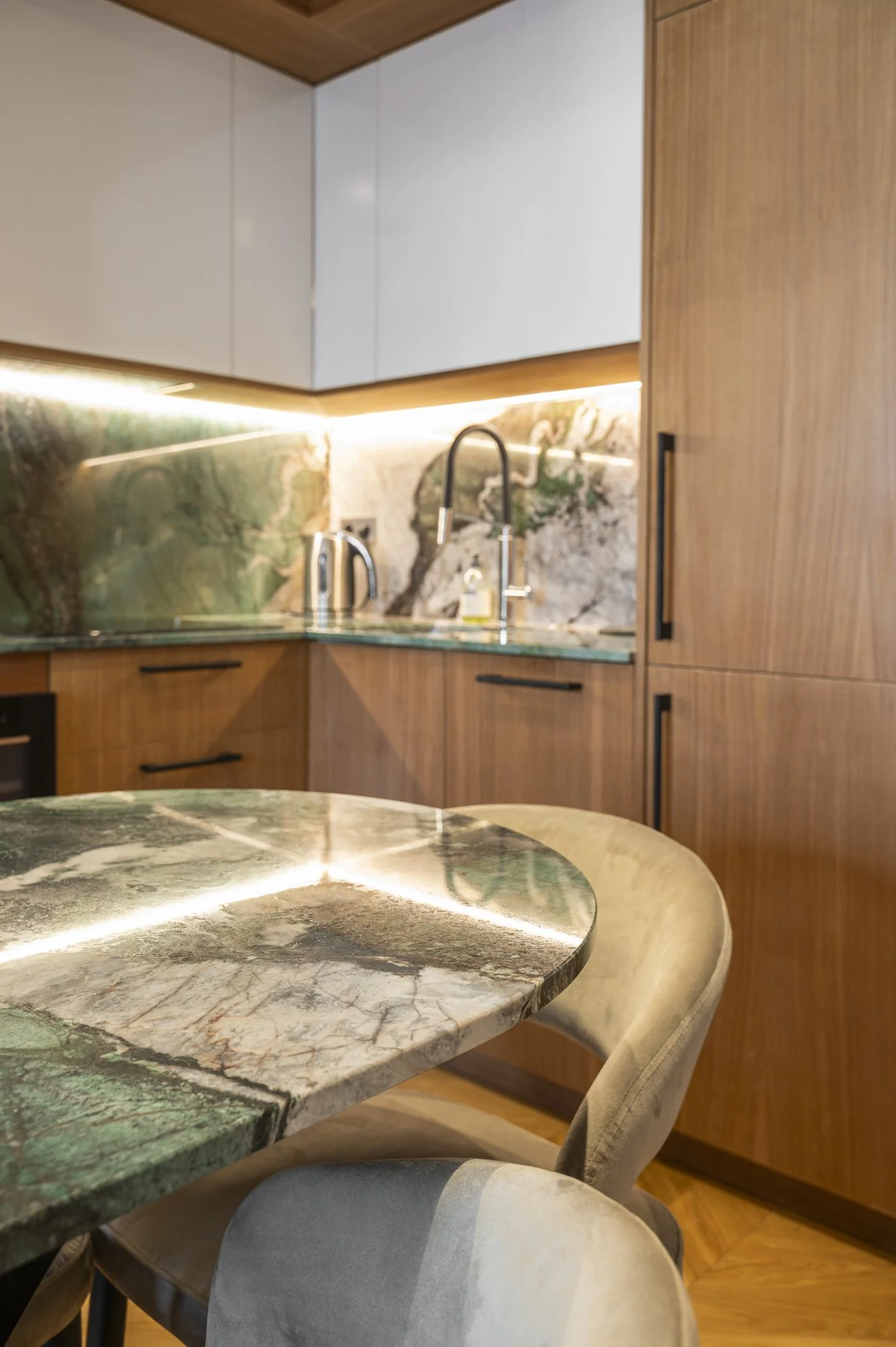 A close-up view of a marble dining table with green, white, and brown veining, accompanied by beige chairs, in a modern kitchen with wooden cabinets and a marble backsplash.
