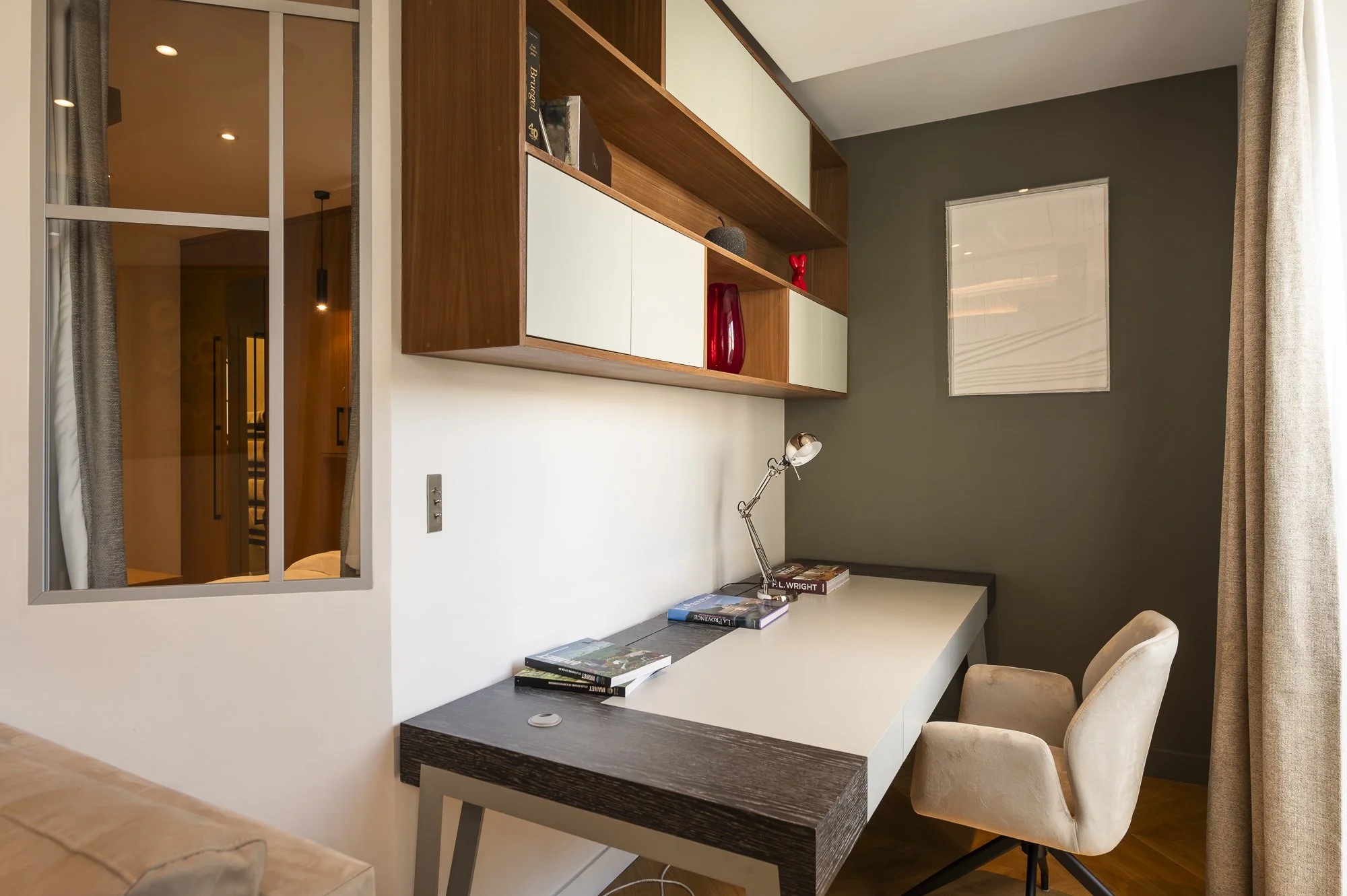 A modern home office with a white and dark wood desk, beige office chair, wall-mounted wooden and white storage cabinets, books, desk lamp, and a window with beige curtains.