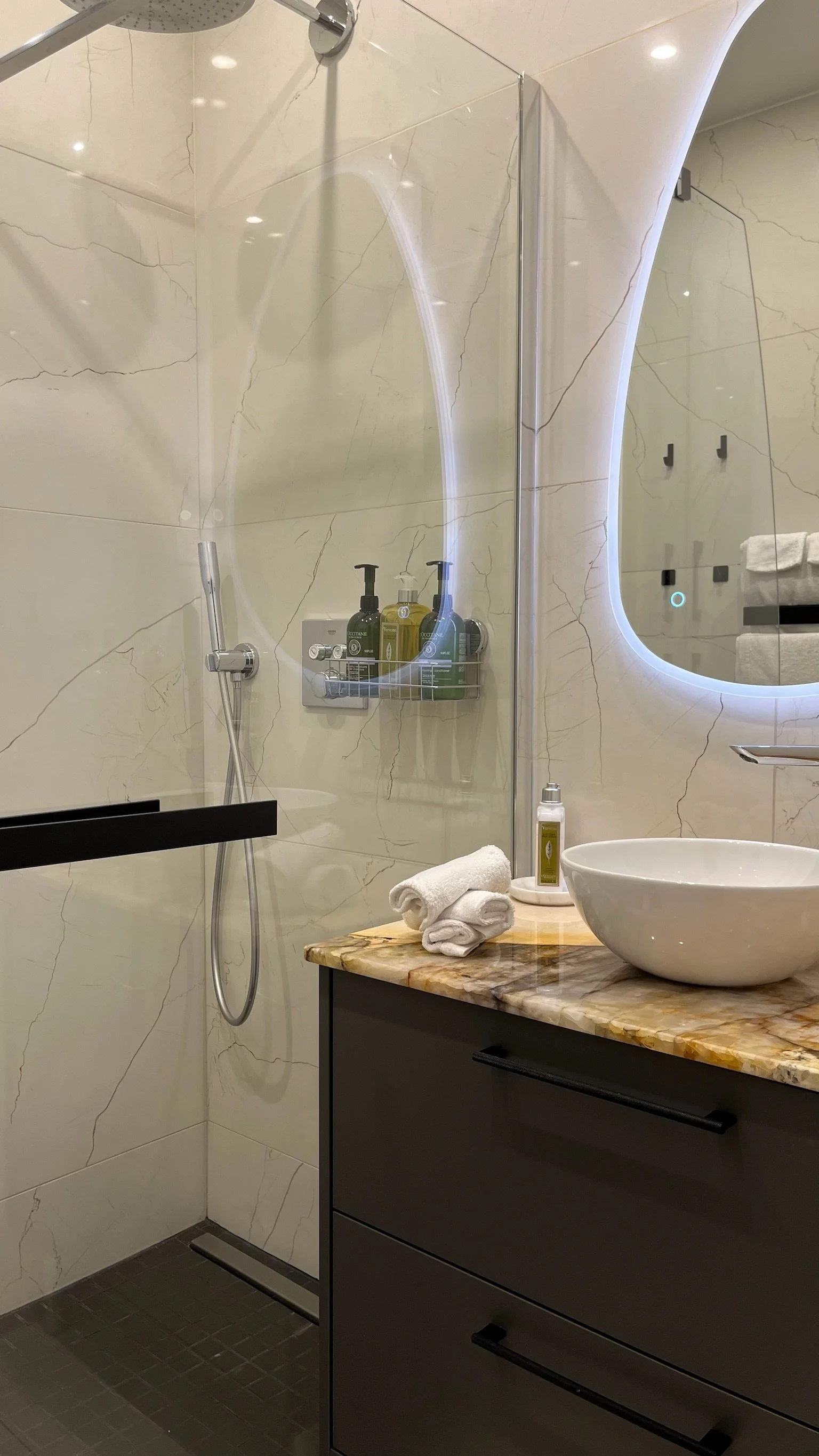 A modern bathroom with a walk-in shower with a glass door, marble walls, a wall-mounted shower head, and a black grab bar. A dark vanity with a round vessel sink, a mirror with backlit LED lighting, a few rolled white towels, and assorted toiletries.