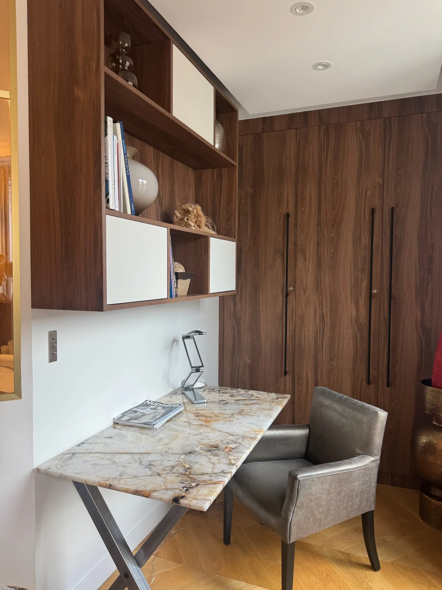 A home office corner with a marble top desk, a gray upholstered chair, wooden wall and shelf unit, and decorative items on the shelves.